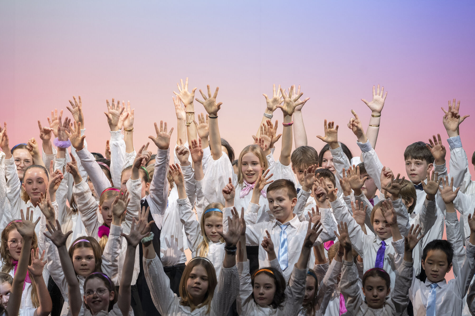 Eine Reihe von Kindern steht auf einer Bühne, singt und hebt die Hände nach oben.