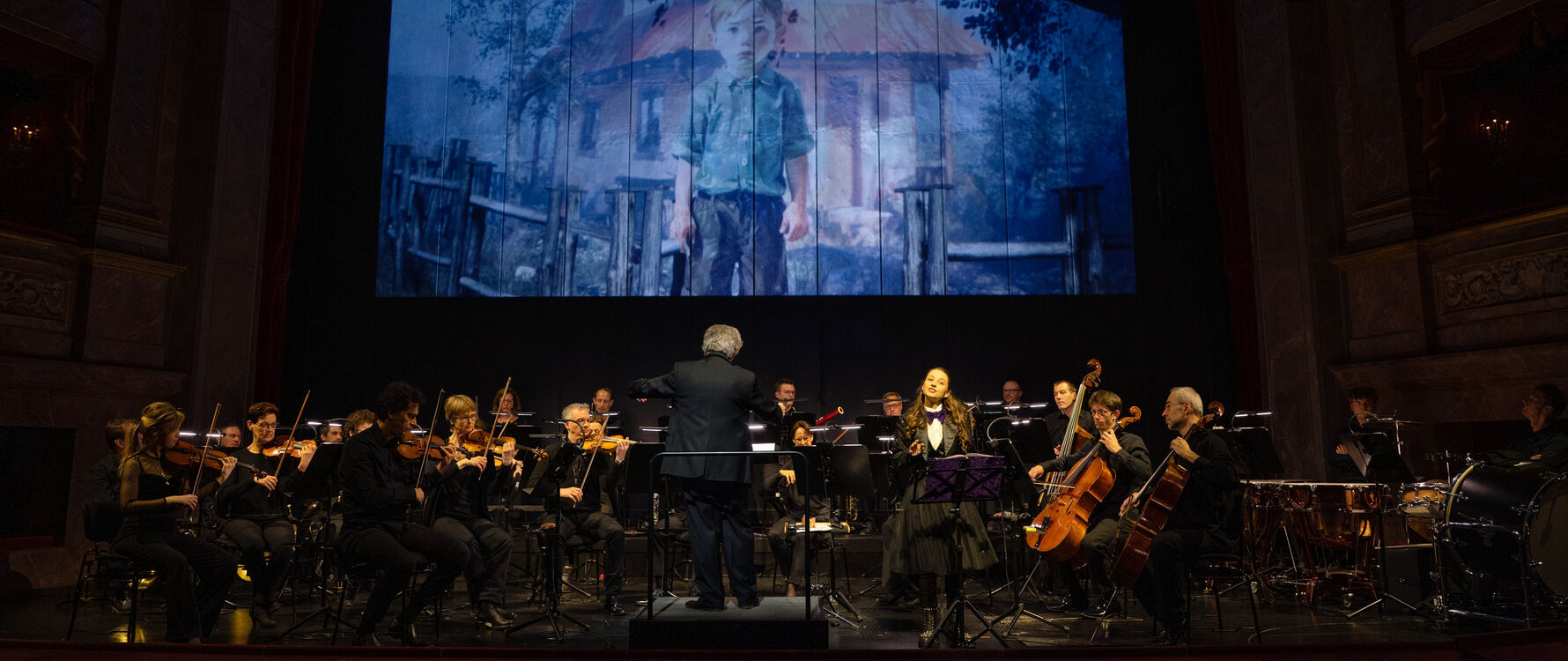 An orchestra sits on a dark stage. In front of them stands a conductor and a narrator who tells the story that can be seen on the screen behind the orchestra.