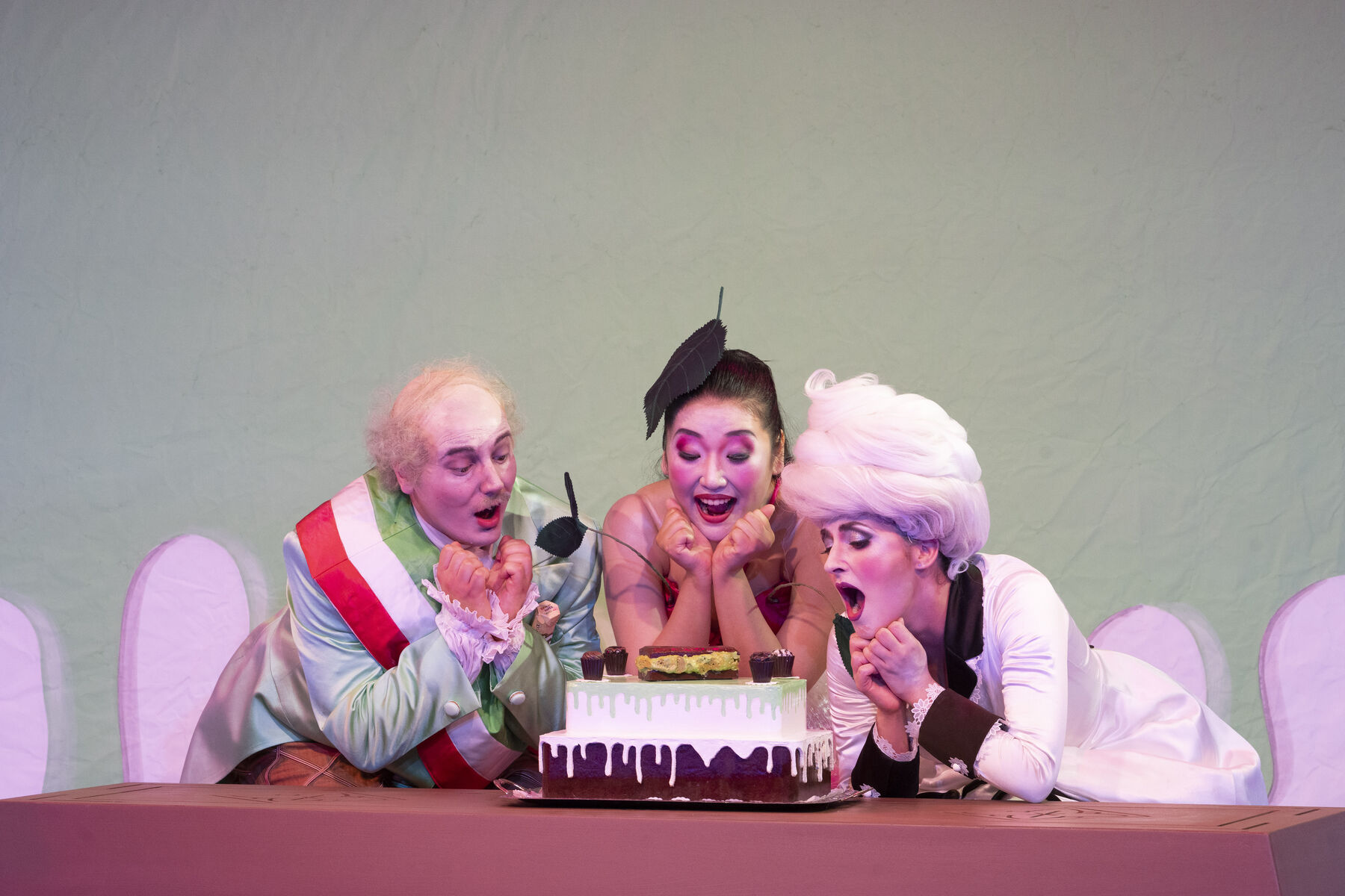 Ein Mann und zwei Frauen beugen sich in einem knallbunten Bühnenbild über einen Kuchen und singen.