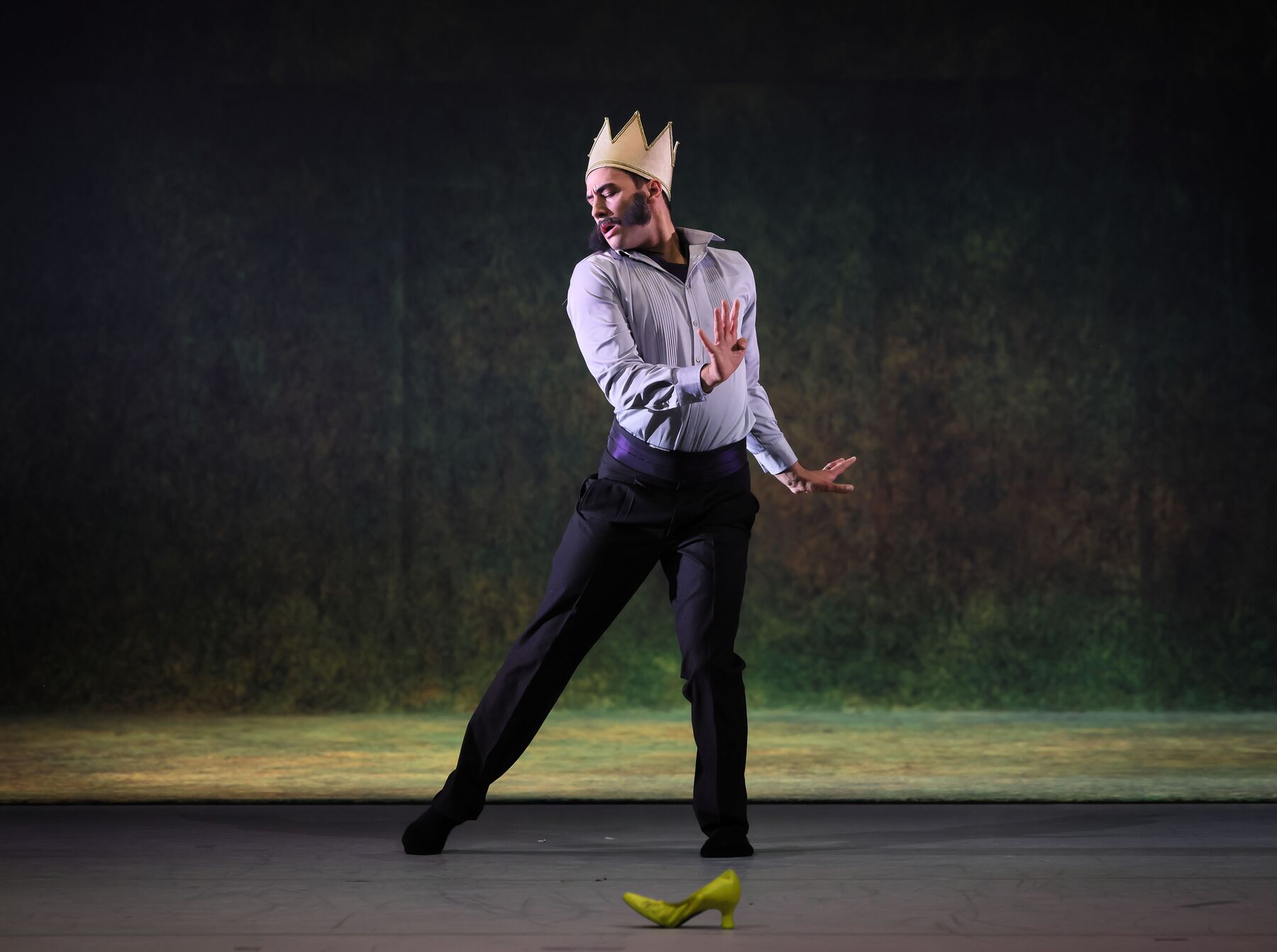 Der König tanzt und trägt dabei eine dunkle Hose und ein hellblaues Hemd. Auf dem Kopf trägt er eine goldene Krone. Vor ihm am Boden steht ein hellgrüner Stöckelschuh. 