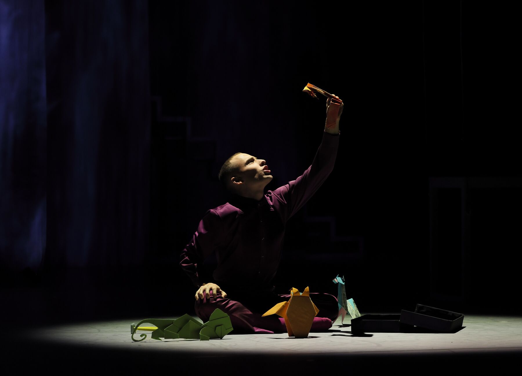 Ein Mann in lilafarbenen Gewand sitzt auf dem Boden und blickt nach oben, während er eine Figurine aus Papier in der linken Hand hält. Um ihn herum liegen weitere Figuren aus buntem Papier.