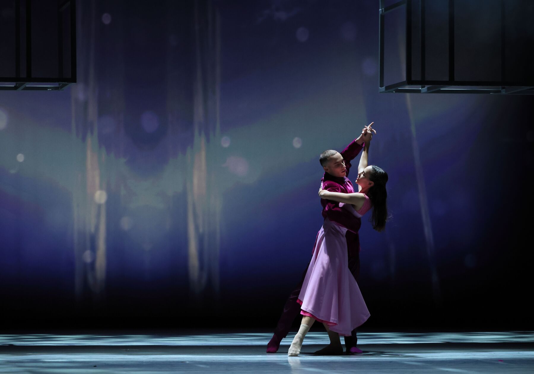 Ein Mann im lilafarbenen Gewand hält eine Frau im rosa-roten Kleid. Sie tanzen und schauen sich an.