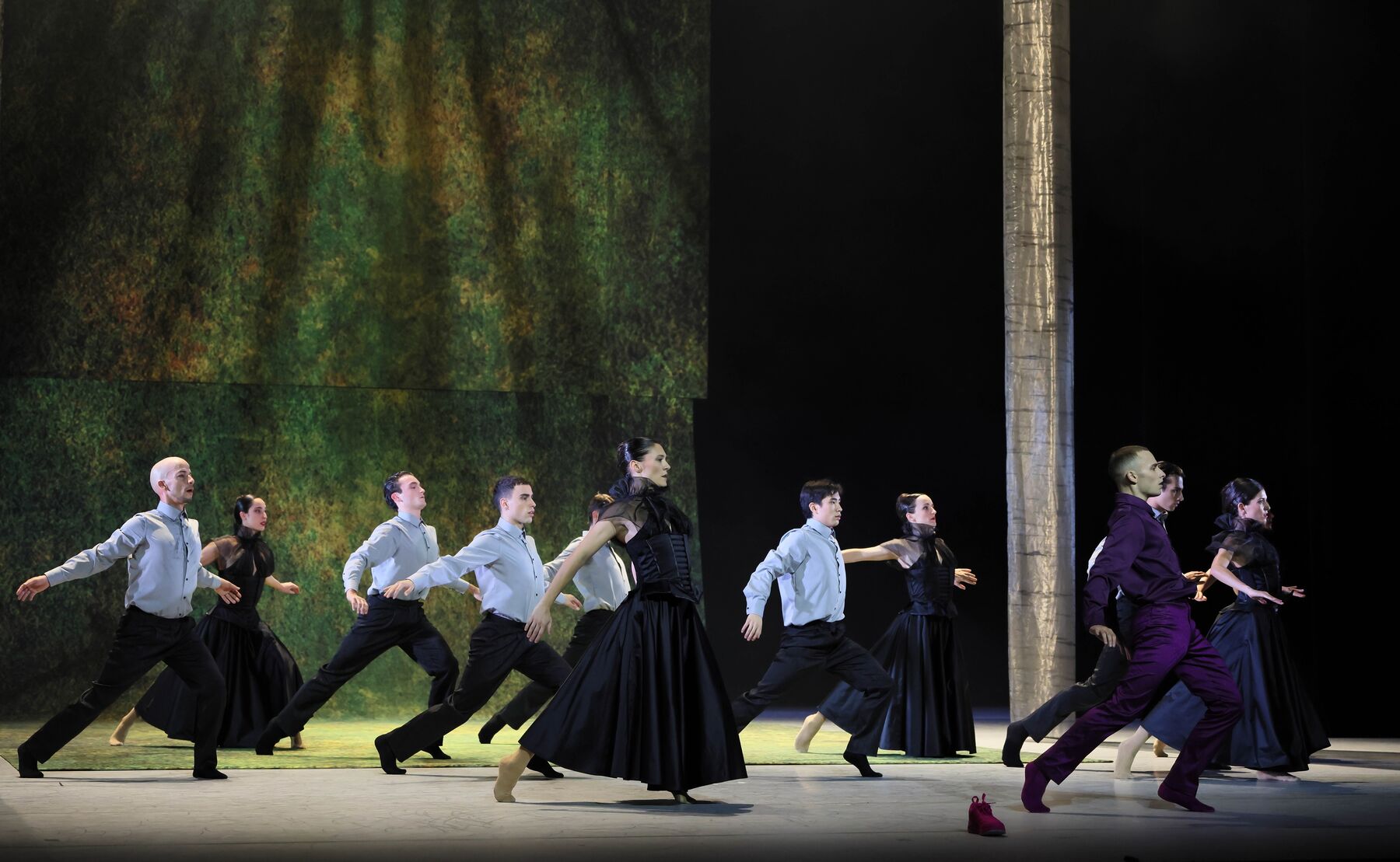 Eine Gruppe von Tänzerinnen und Tänzer in schwarzen Kleider und grauen Hemden mit schwarzen Hosen, strecken ihre Arme zur Seite und machen alle den gleichen Schritt zur Seite.