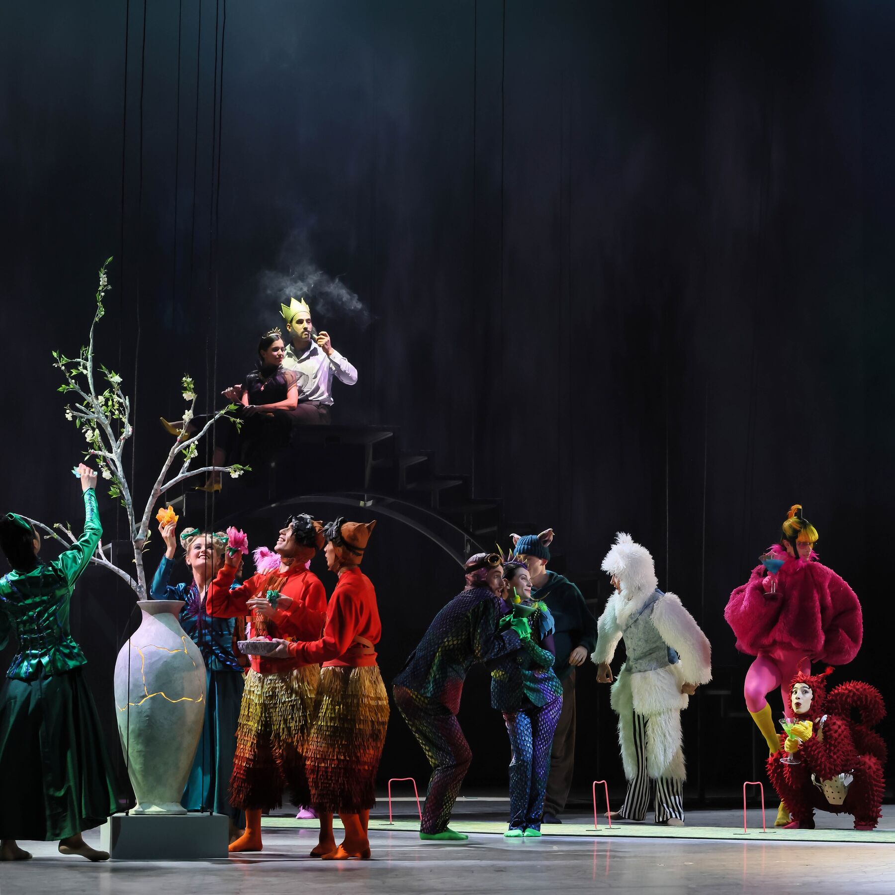 Eine Gruppe von Personen in unterschiedlichen Tierkostümen ist zu sehen. Ein paar von ihnen stehen um eine weiße Vase aus der ein Zweig mit grünen Knospen emporwächst. Im Hintergrund sitzt ein Mann mit Krone auf einer Treppe. Neben ihm sitzt eine Frau mit Diadem und schwarzem Kleid.