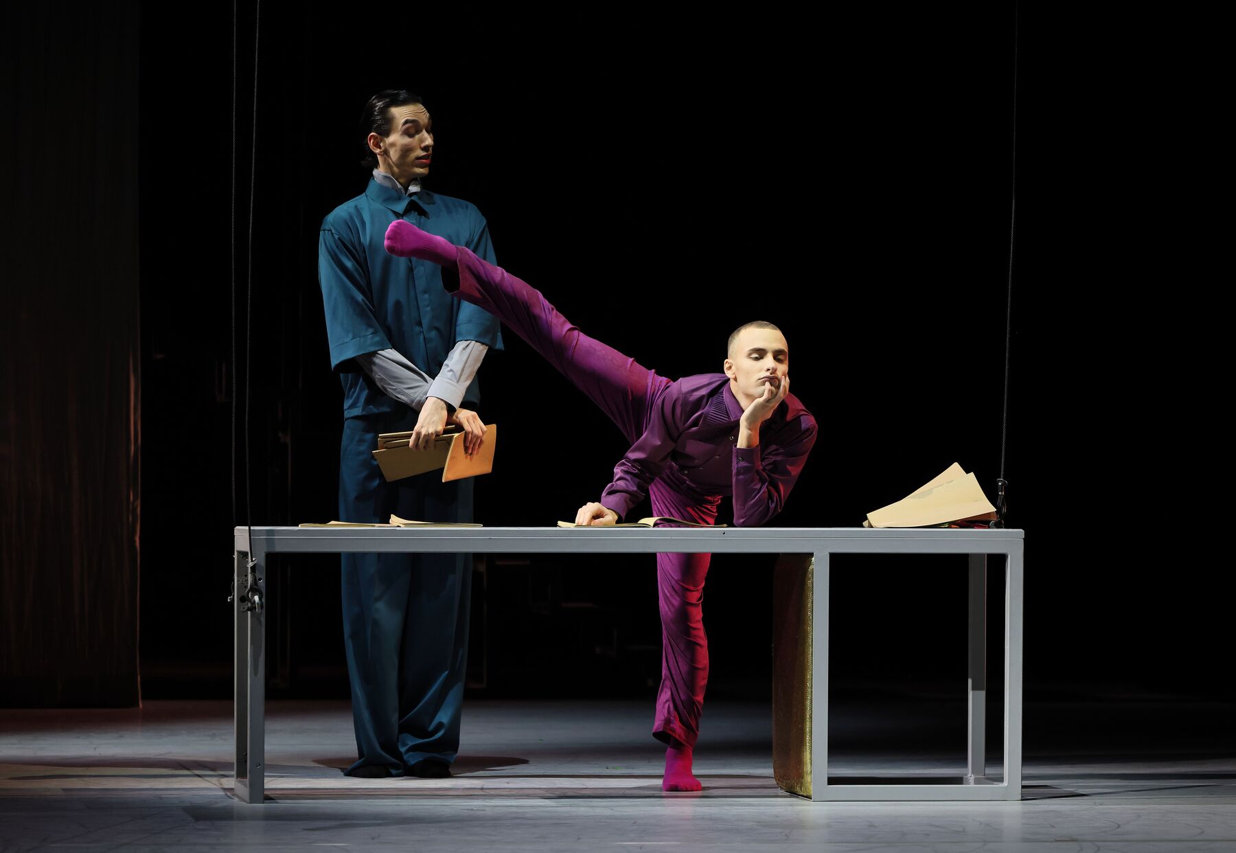 Der Prinz stützt sich auf den Tisch und zieht eine Grimasse. Hinter ihm steht der Prinzenerzieher und lacht.