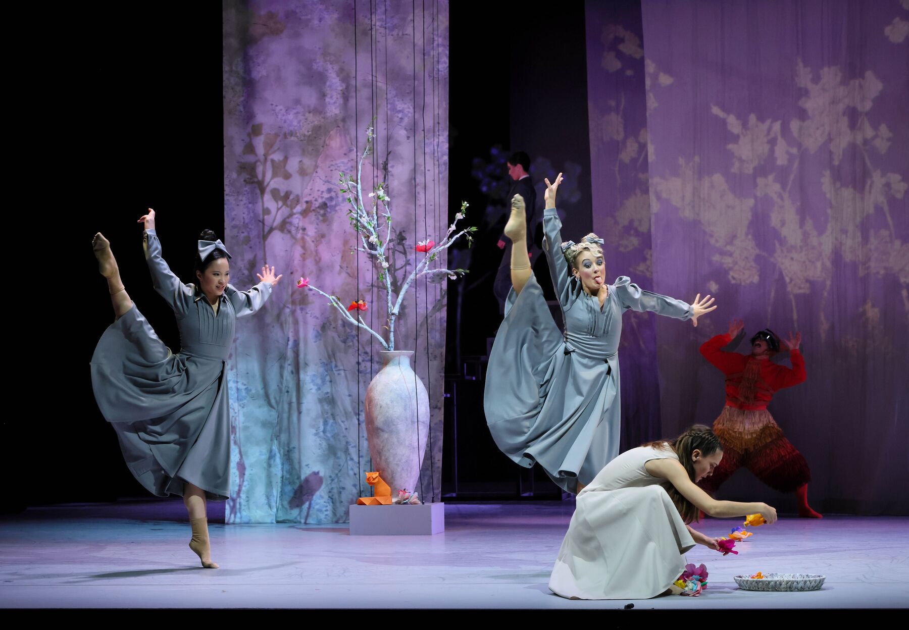 Aschenbrödels Stiefschwestern tanzen um sie herum und ziehen Grimassen. Aschenbrödel kniet auf dem Boden und hält Papierblumen in der Hand.