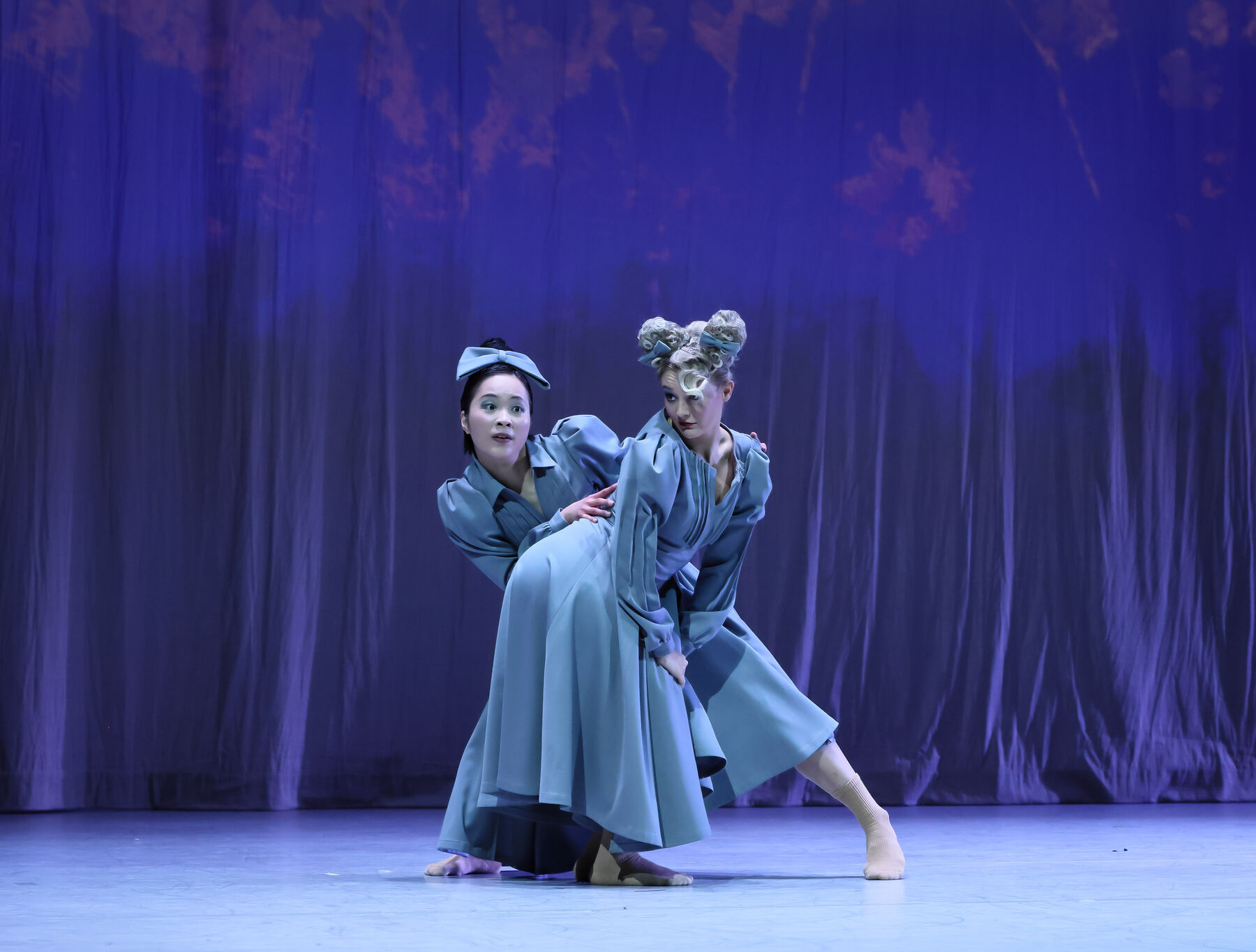 Zwei Tänzerinnen in blauen Kleidern und blauen Fliegen in den Haaren stehen hintereinander, wobei die eine links an der anderen vorbeiblickt. 