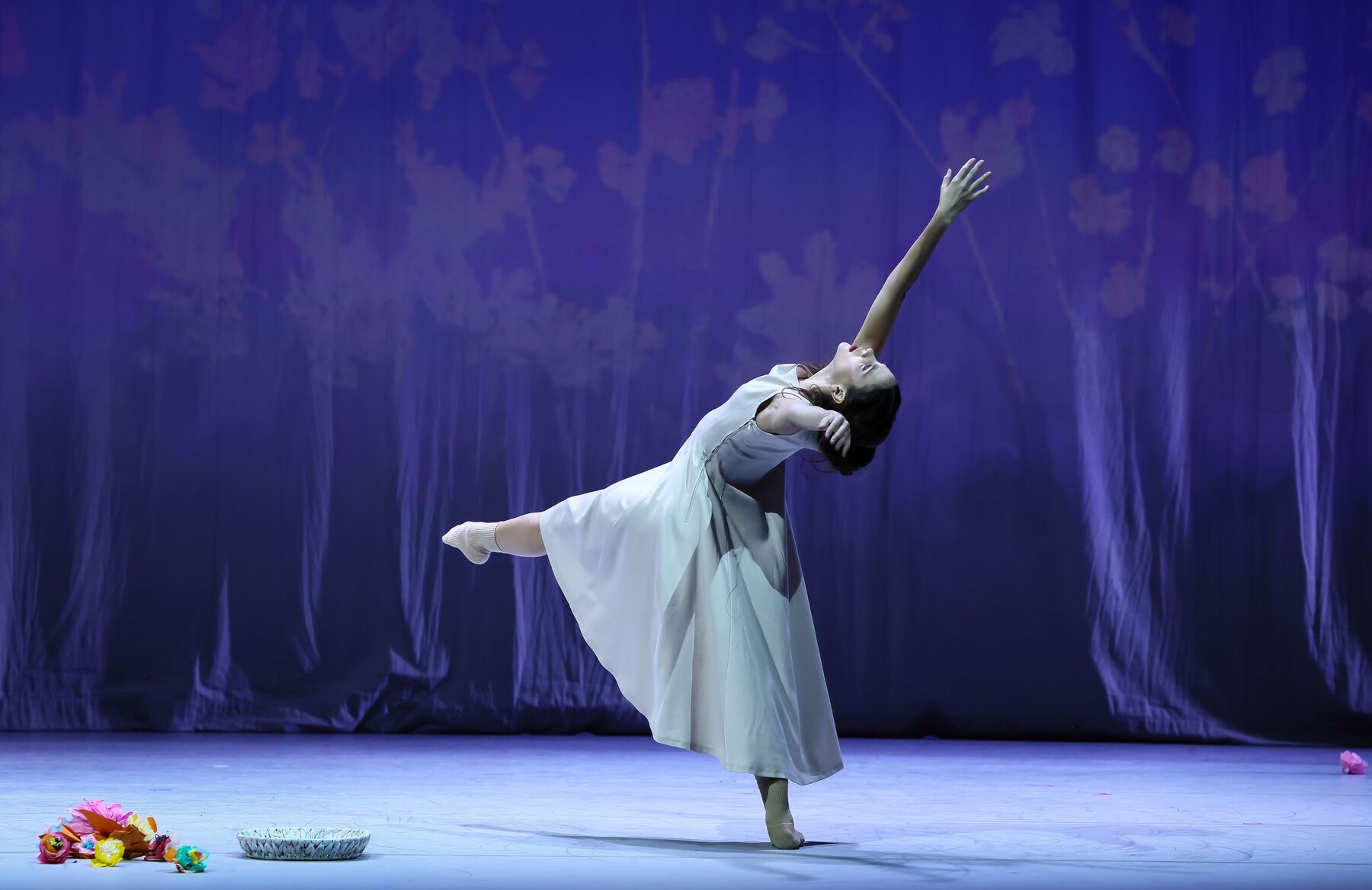 Eine Frau im weißen Kleid steht auf einem Bein, während sie das andere nach vorne ausstreckt. Ihre Hände und ihren Oberkörper wiegt sie nach hinten. Auf dem Boden vor ihr liegt ein weißer Korb und daneben Blumen.