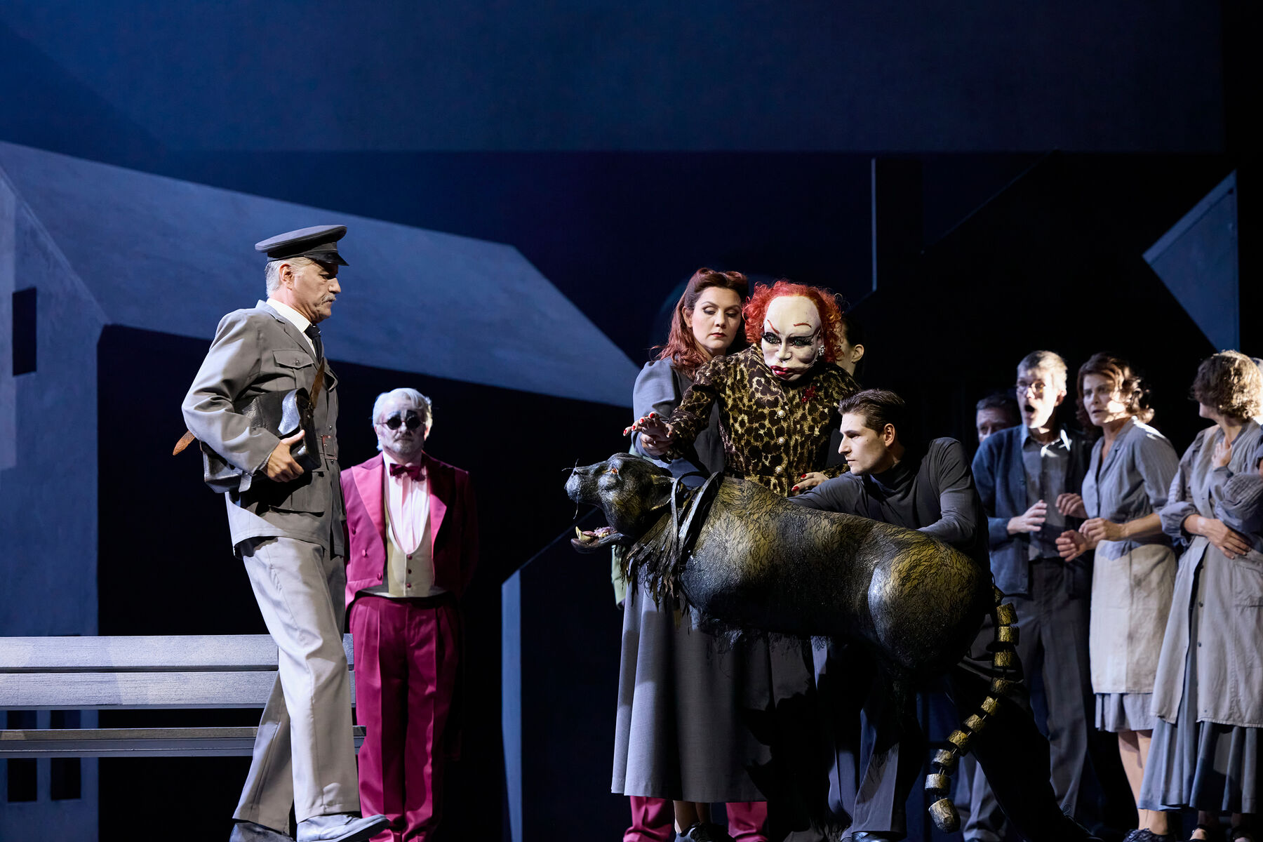 ein ältere Mann in einer grauen Zugführeruniform steht gegenüber von dem schwarzen Panther. Eine Frau mit roten Haaren und einem grauen Kleid und eine lebensgroße Puppe von einer älteren, rothaarigen, schickgekleideten aber entstellten Dame, halten den Panther zurück. Im Hintergrund steht ein Mann mit rotem Frack und Sonnenbrille und ein paar Menschen in grauer Alltagskleidung.