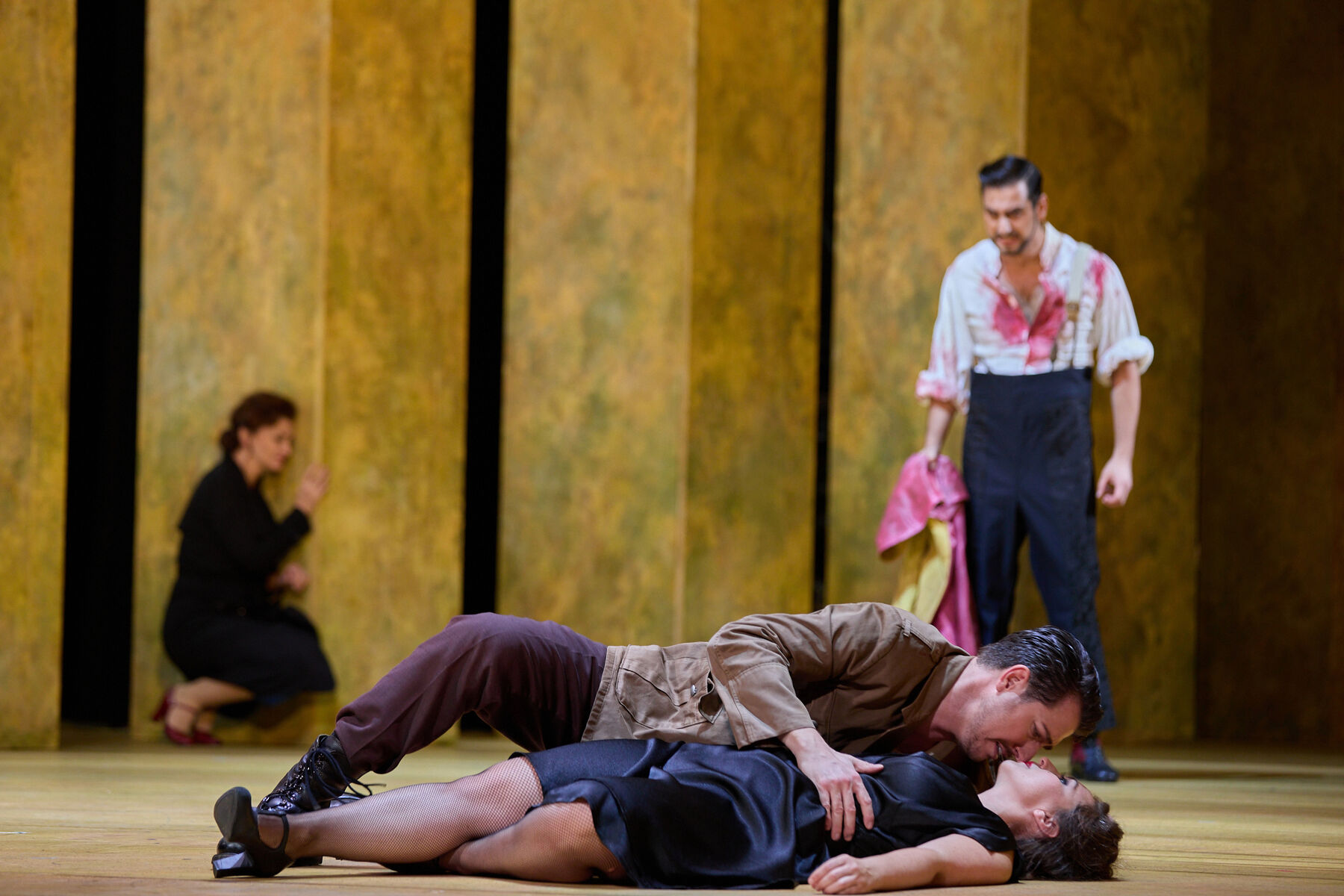 Carmen lies dead on the floor while Don José bends over her, looking worried. In the background, Micaëla kneels against a wall. On the right stands Escamillo, covered in blood and furious.