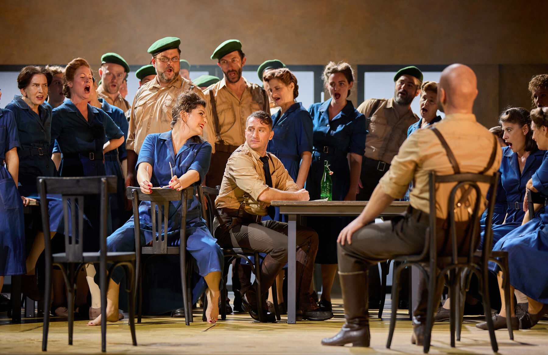 Don José and Carmen are sitting on chairs. A group of women in blue overalls and men in uniform are standing around them.