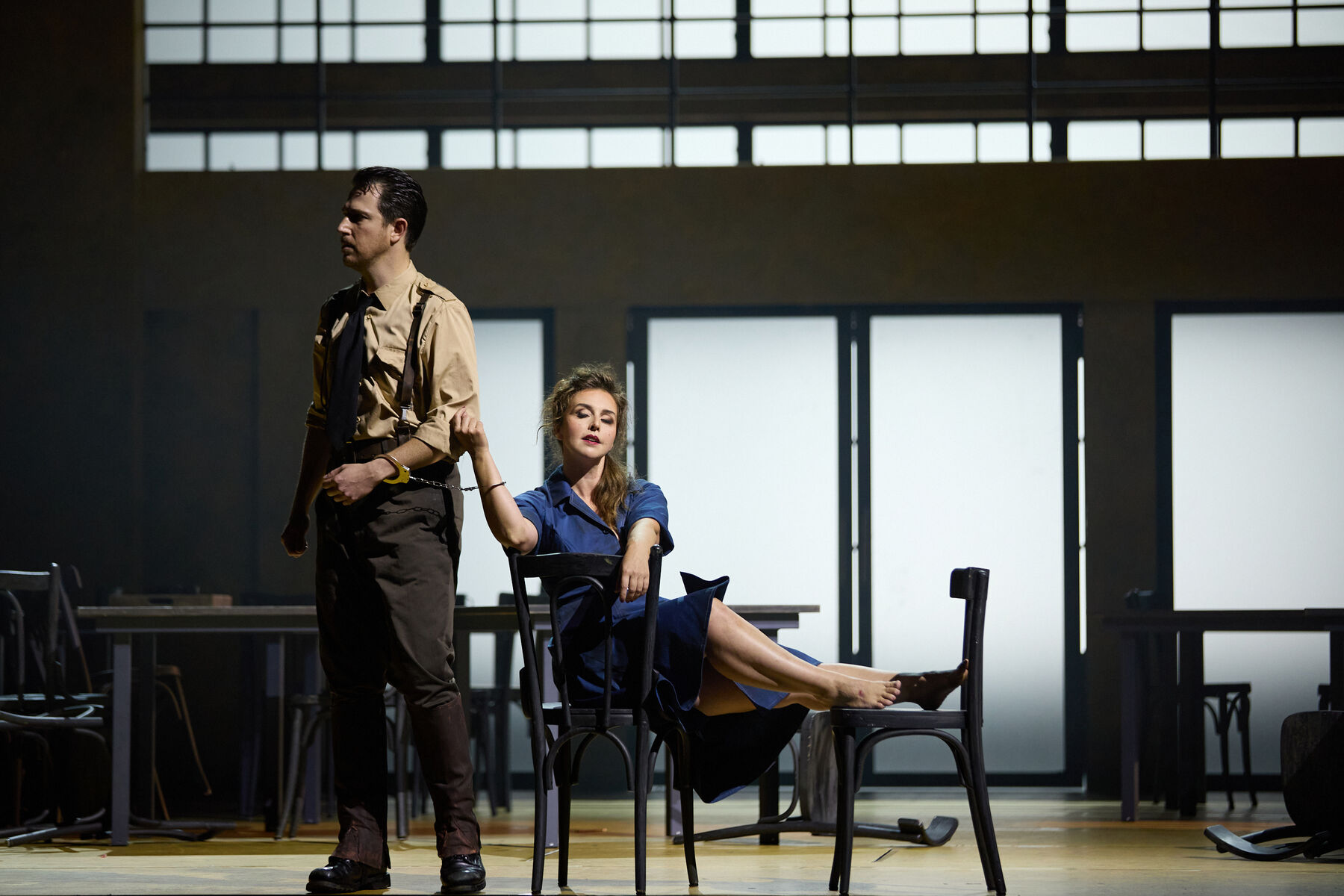 Carmen, reclining on a chair, is chained to Don José by handcuffs.