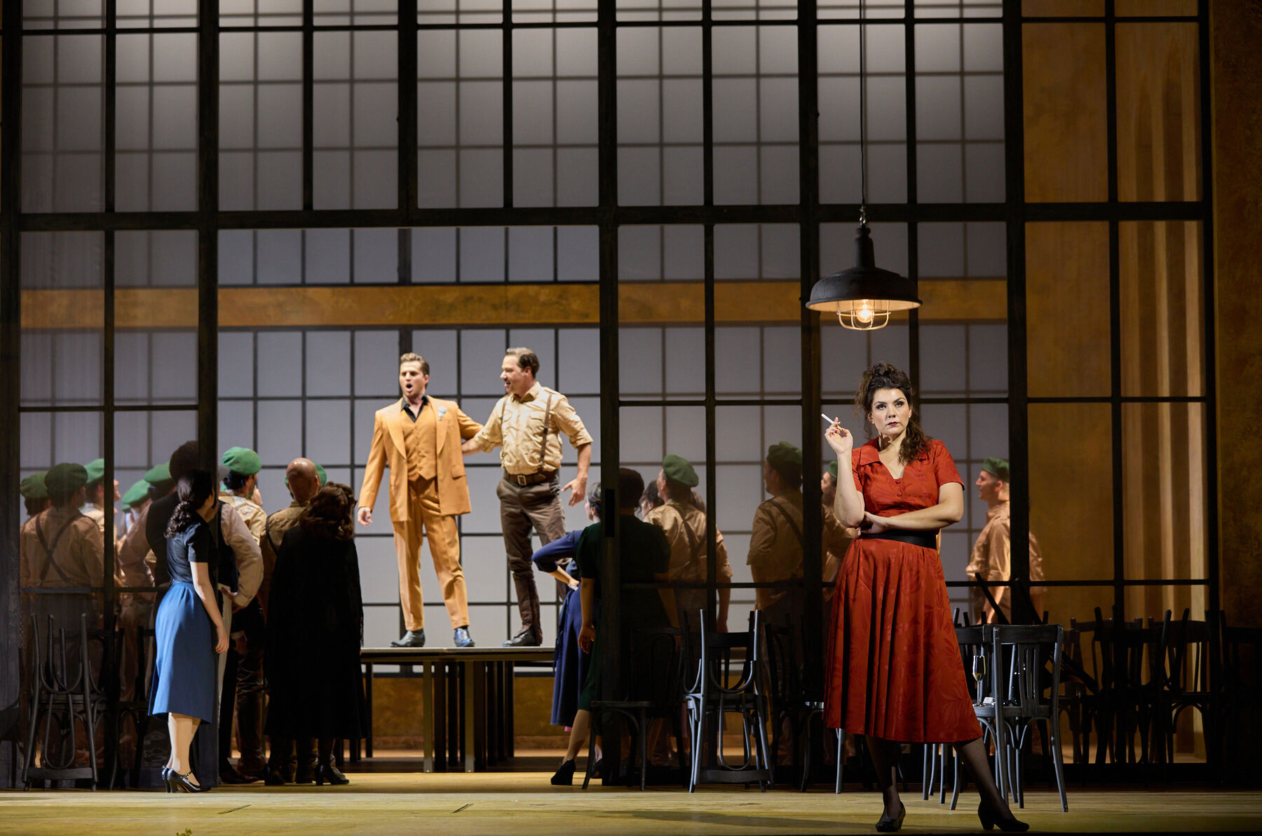 Two men are standing on a table in the background. They are surrounded by a group of people. In the foreground, a woman in a red dress is smoking a cigarette.