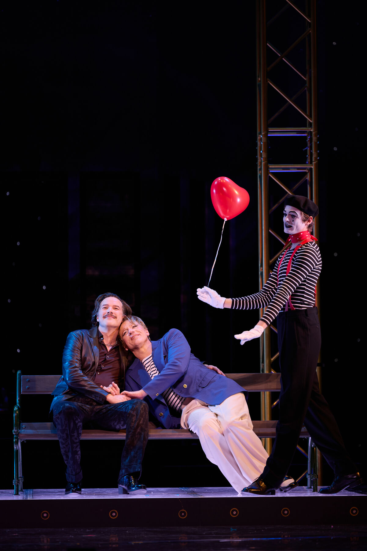 Albin und Georges sitzen kuschelnd auf einer Bank. Daneben hält ein Pantomime einen roten Herzballon. 