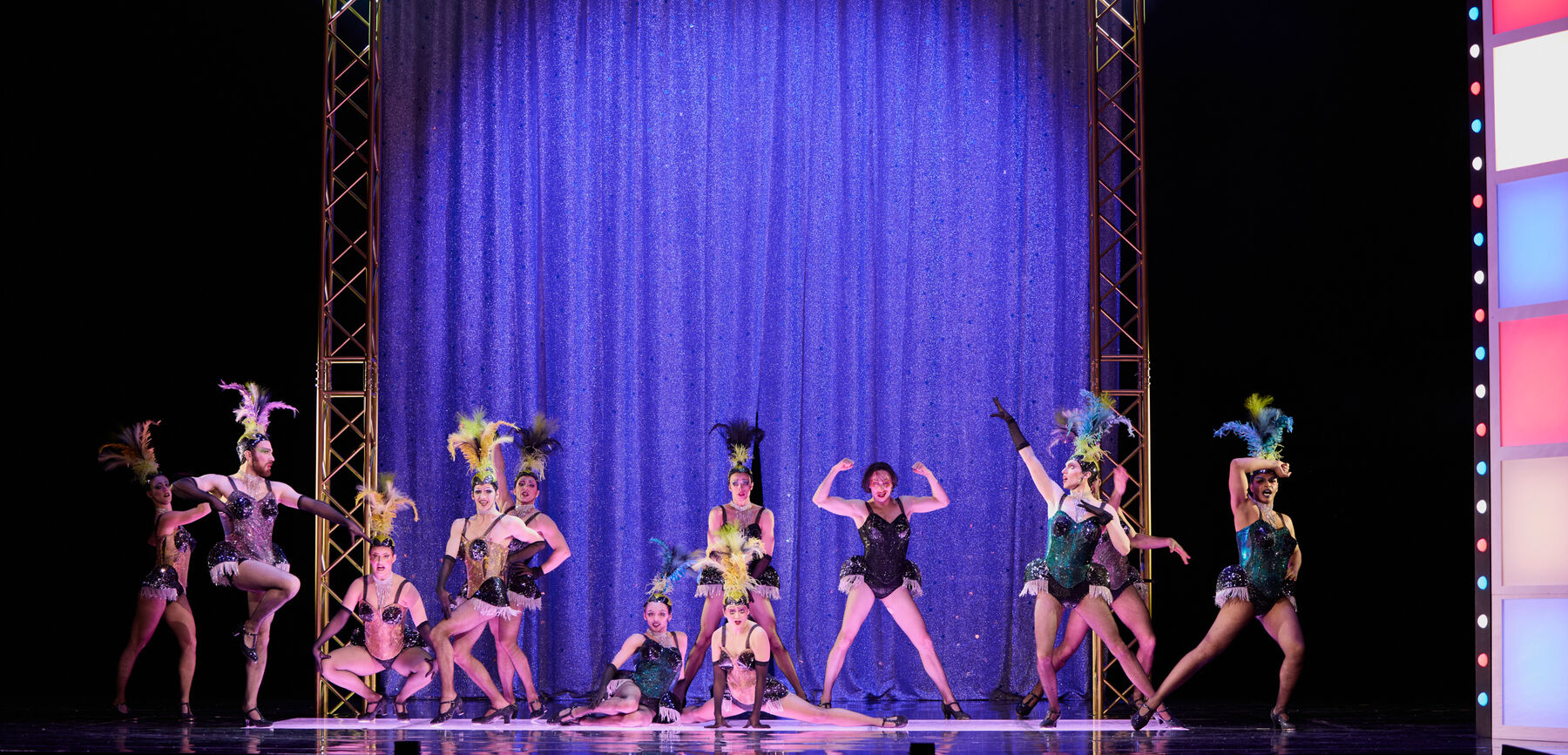 Eine Gruppe von Tänzern in Burlesque-Kostümen posiert und performt vor einem lila-glitzernden Vorhang.