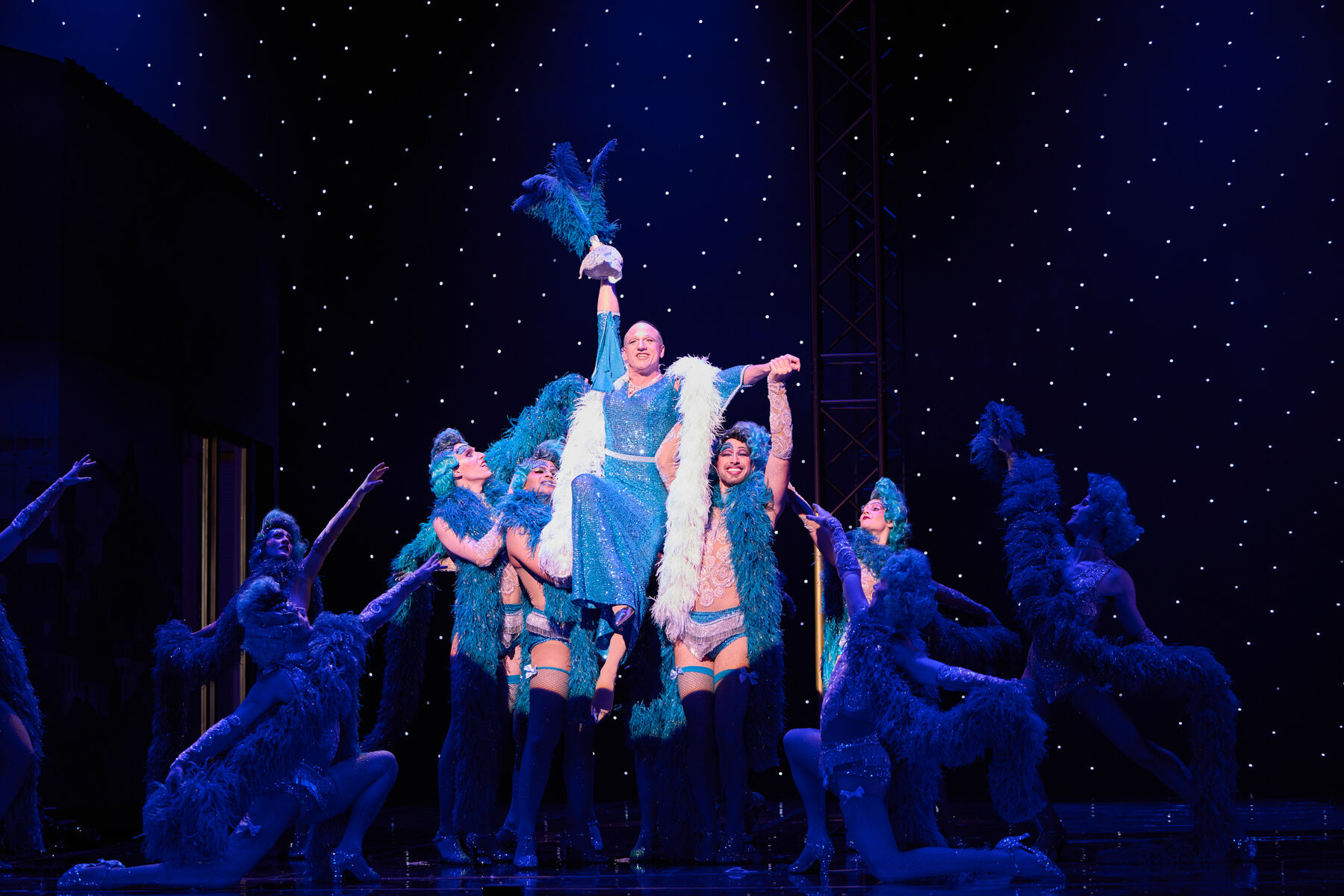 Albin wird von Tänzern hochgehoben, während er im langen blauen Glitzerkleid performt.