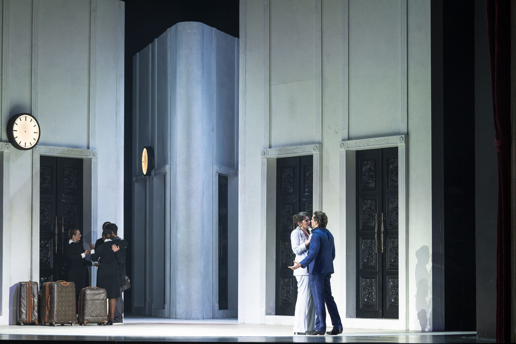 Don Giovanni and Donna Elvira are facing each other and appear to be engaged in conversation. In the background, other people can be seen pushing several suitcases.