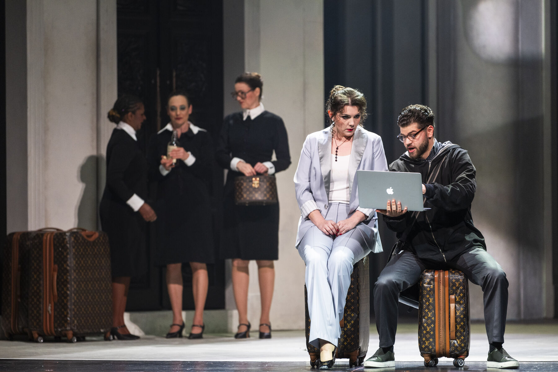 Donna Elvira and Leporello are sitting on suitcases. Leporello shows Donna Elvira something on a laptop. In the background, three formally dressed ladies and more suitcases can be seen.