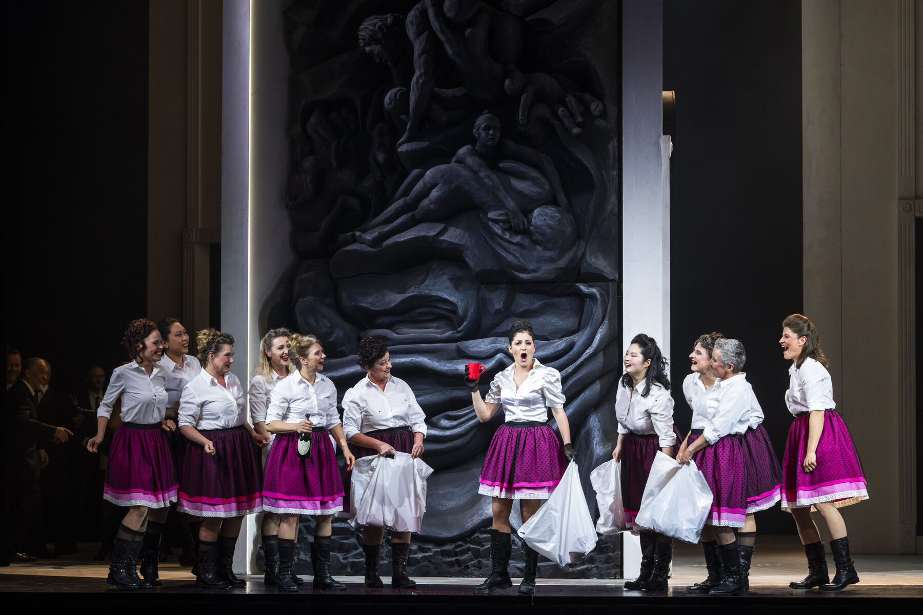 Eine Gruppe von lachenden Frauen in weißem Oberteil und lila-rosa Röcken stehen um eine Frau herum, die dasselbe trägt. Sie hält eine rote Kaffeetasse und ein Kissen in der anderen Hand und singt nach vorne. Im Hintergrund ist ein dunkelgraues weiß-umrahmtes Steinbild zu sehen, das ineinander verschmelzende Körper zeigt.