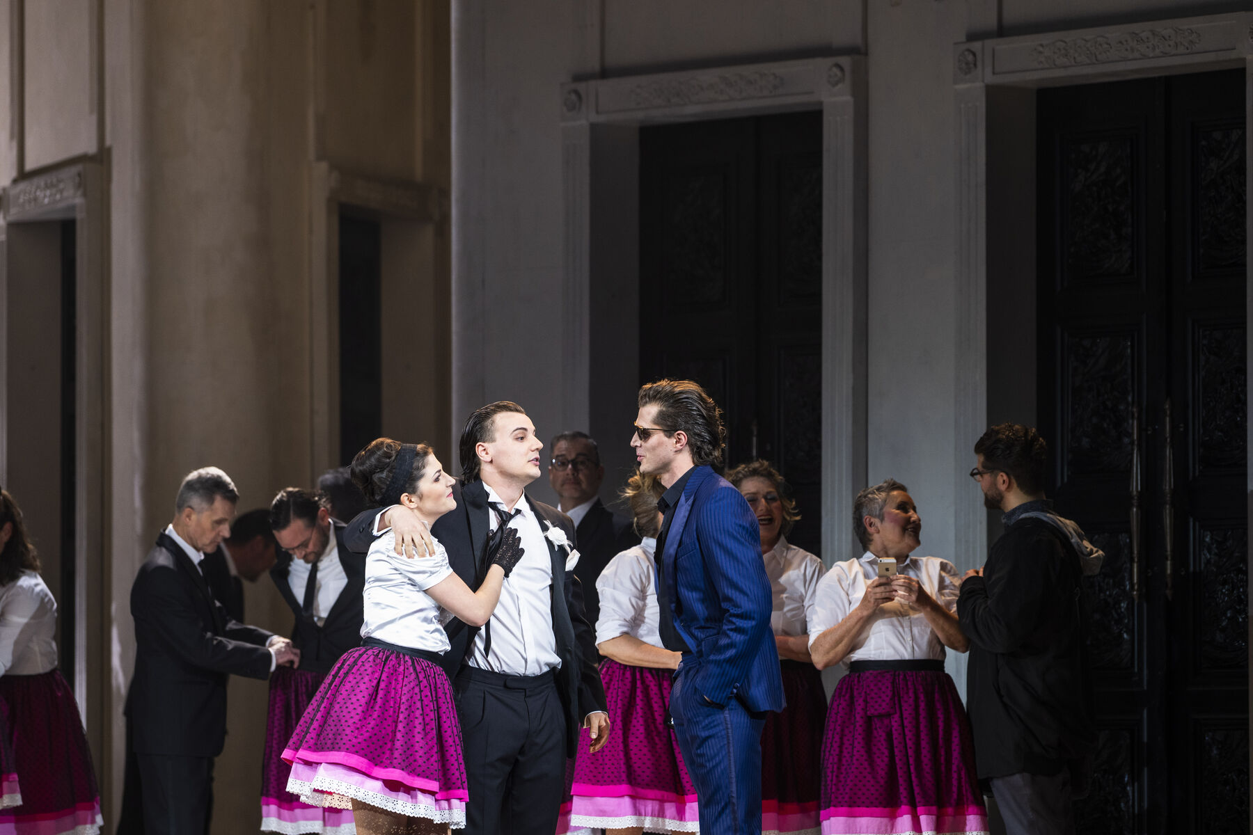 Masetto has his arm around Zerlina and is talking to Don Giovanni, who is standing in front of them wearing a blue suit and sunglasses. In the background are women and men dressed in the same clothes as Masetto and Zerlina.