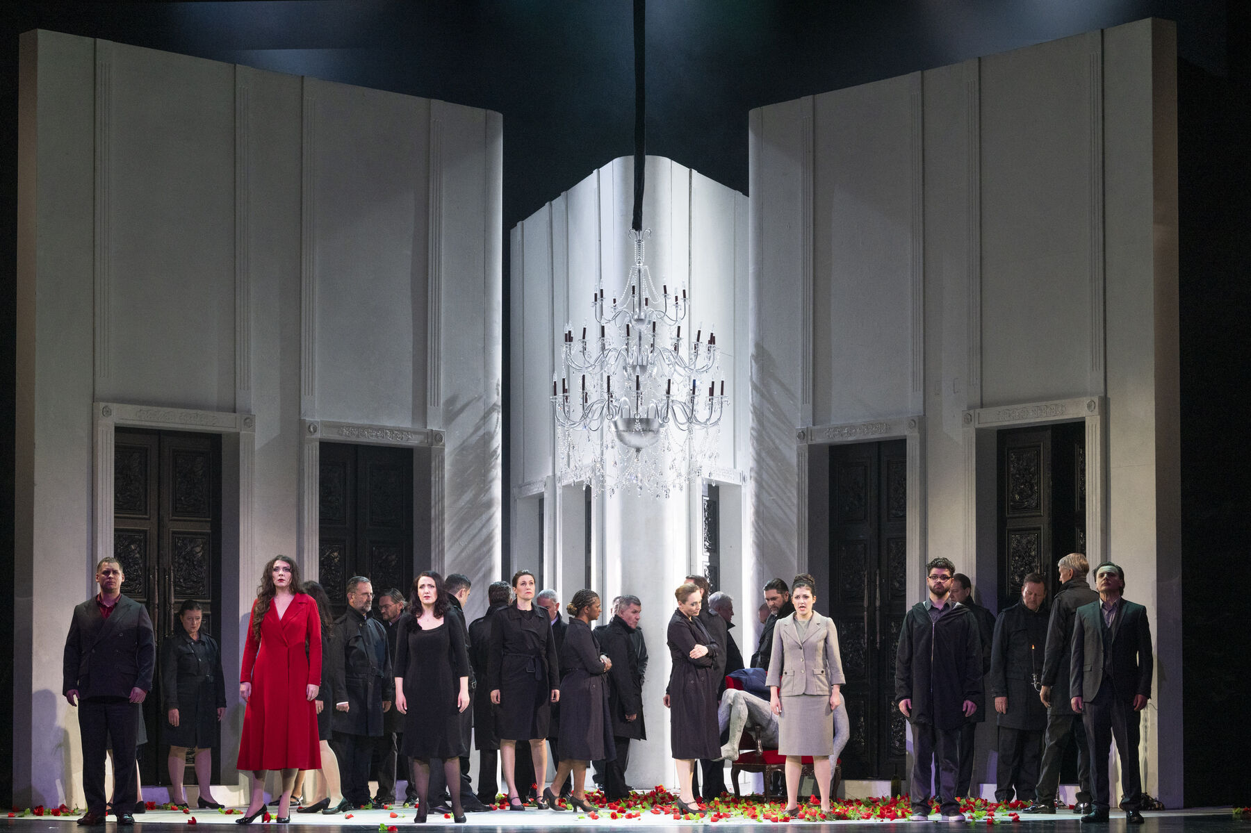 A group of people dressed in different clothes are scattered around the room. A chandelier hangs in the middle of the room and roses are scattered on the floor.