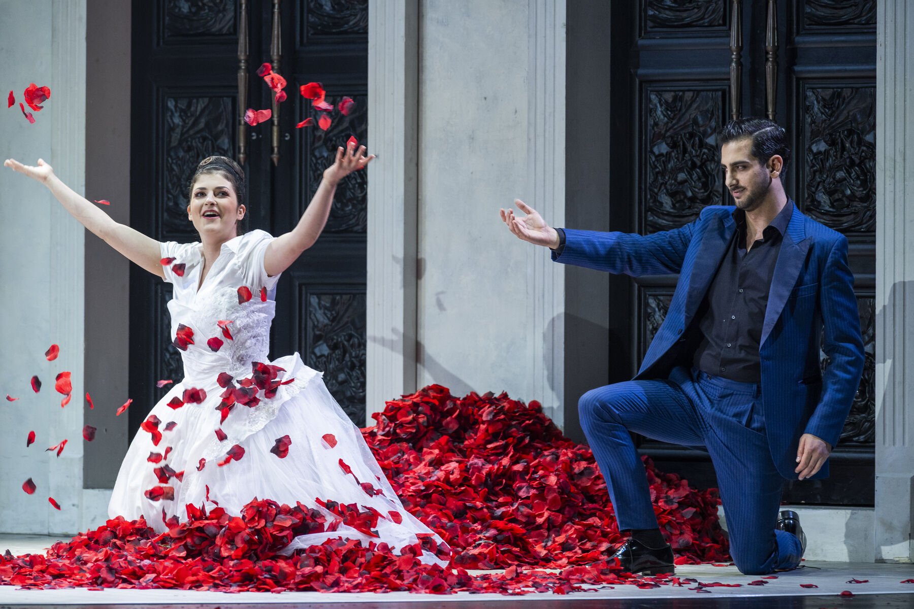 Zerlina, wearing a wedding dress, kneels in a pile of rose petals and throws them into the air. Don Giovanni, also kneeling, points at Zerlina.
