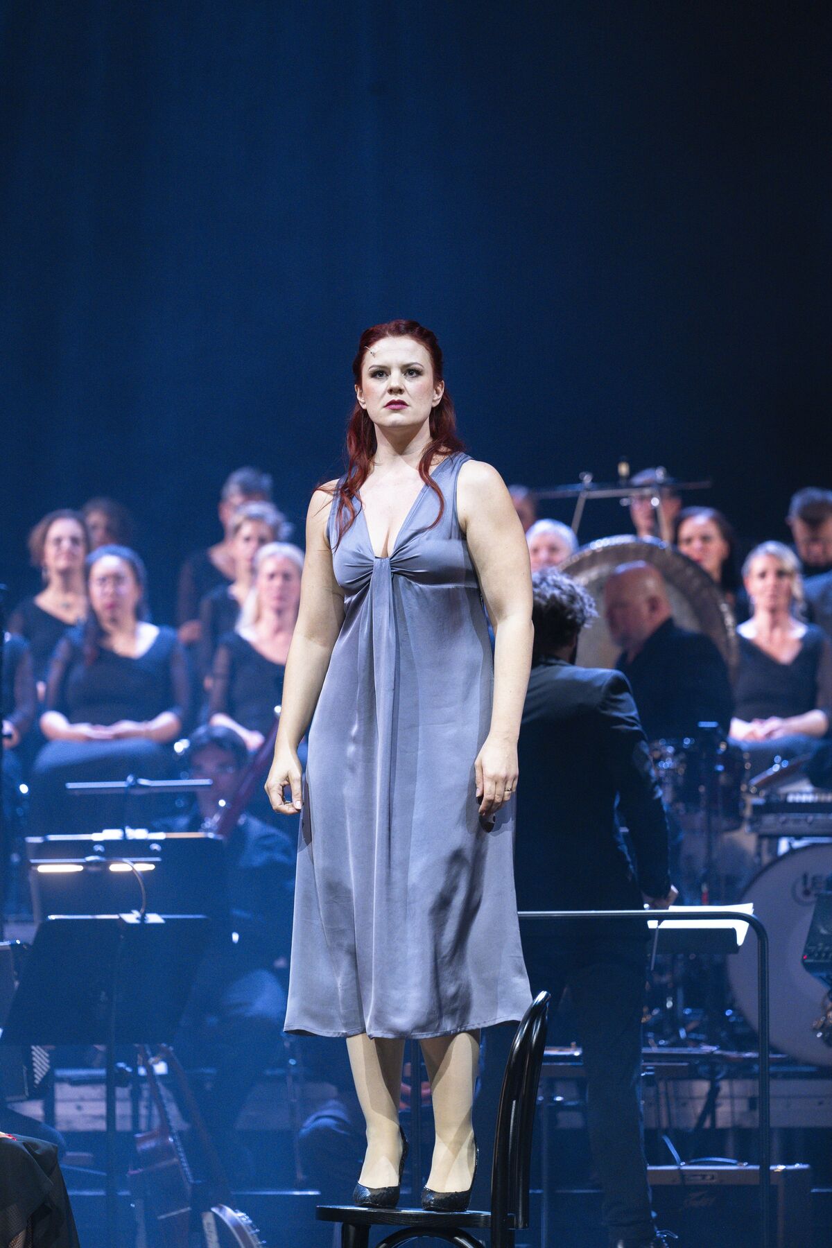 eine Frau in einem blauen Kleid steht auf einem Stuhl. Im Hintergrund ist ein Orchester und ein Chor.