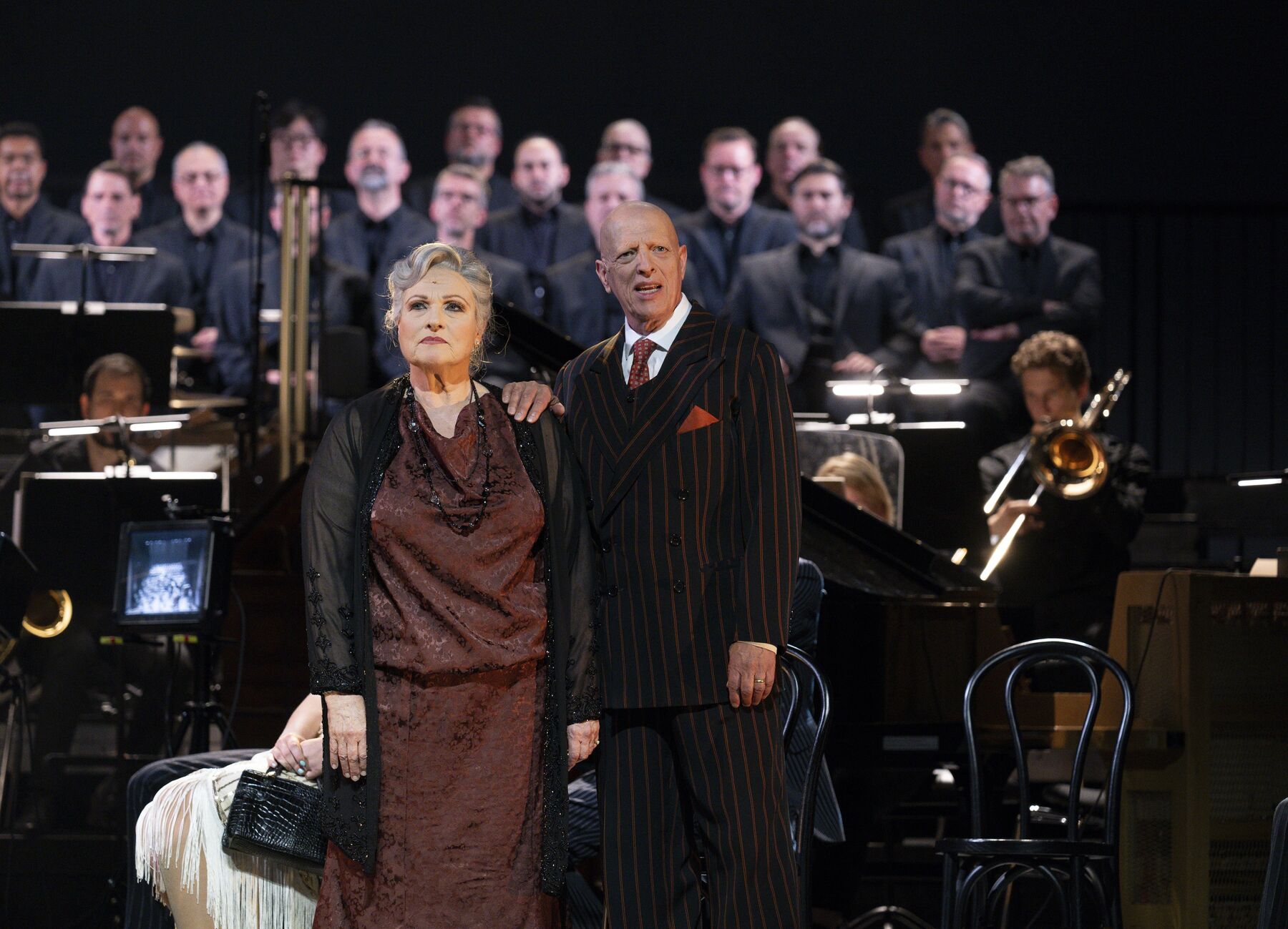 eine ältere Dame in einem roten Kleid und ein älterer Mann in einem schwarz roten Anzug stehen nebeneinander. Im Hintergrund ist ein Chor und ein Orchester.