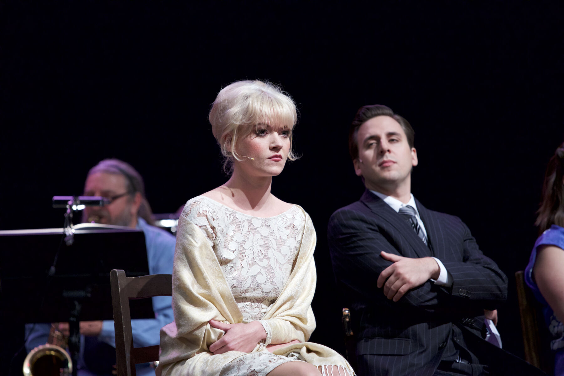 A woman and a man are sitting in front of a dark background. He is looking at her, she is staring intently into the distance.