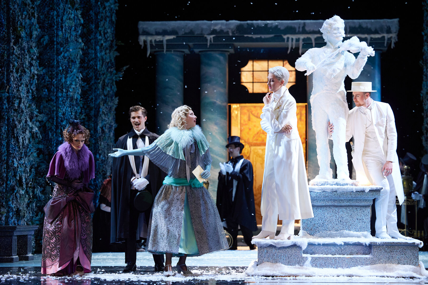 Drei Personen in Ballkleidung stehen neben einer weißen  Statue vor der zwei ganz in weiß gekleidete Menschen stehen.