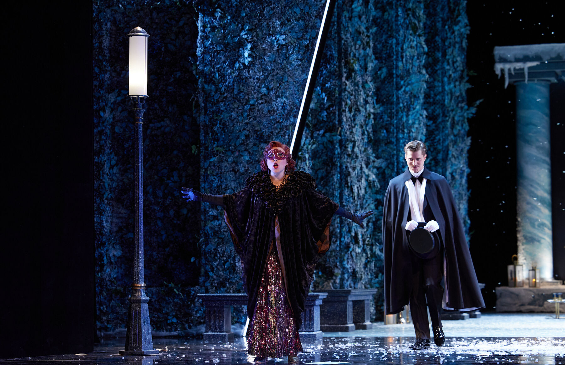 Eine Frau in Ballkleid mit Maske steht neben einer Straßenlaterne. Ihr folgt ein Mann mit Anzug und Hut.