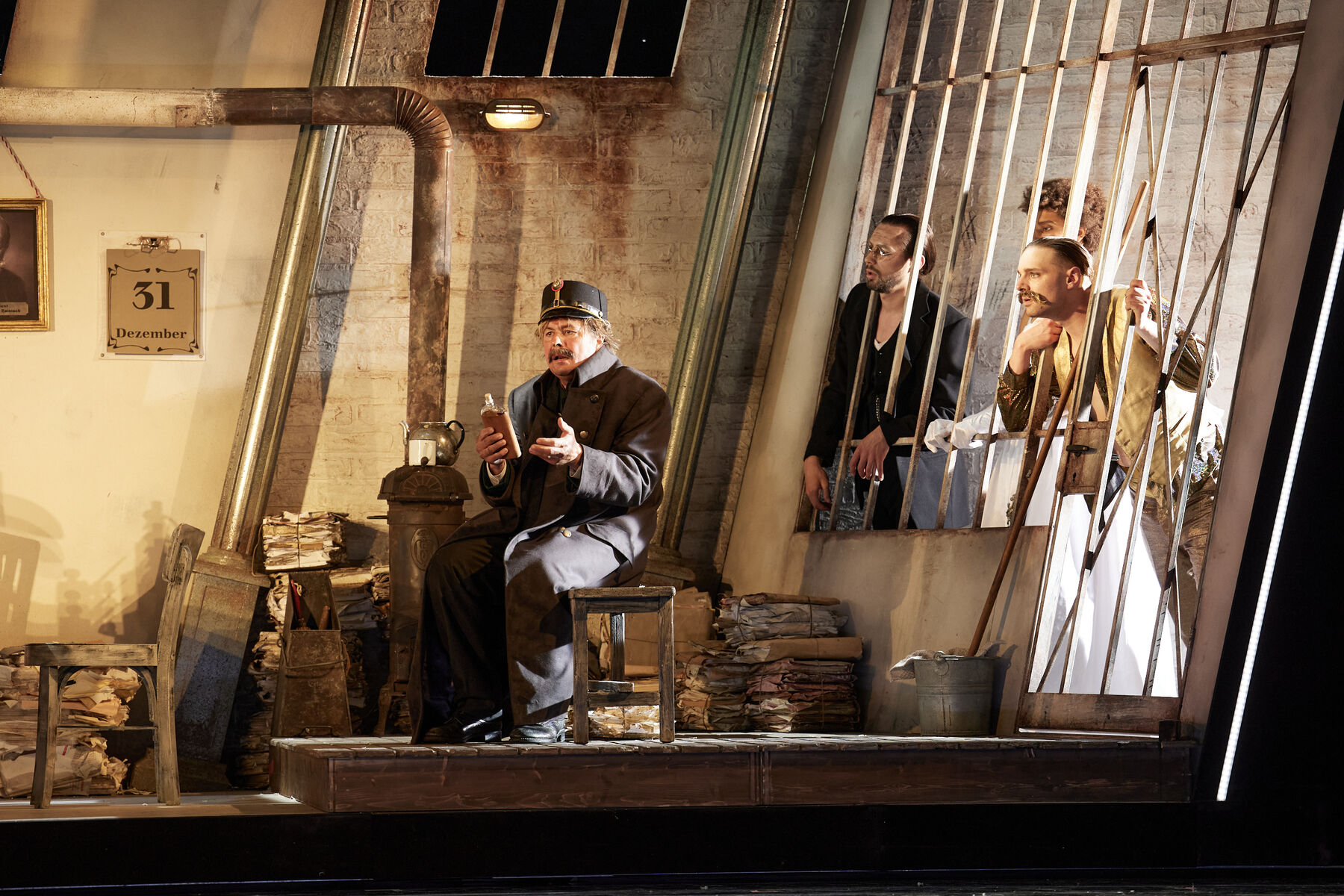 Ein Mann in Uniform sitzt vor einer Gefängniszelle und trinkt aus einem Flachmann. Hinter ihm sind drei Gefangene zu sehen, die ihre Köpfe durch die Gefängnisgitter strecken und ihn anschauen.