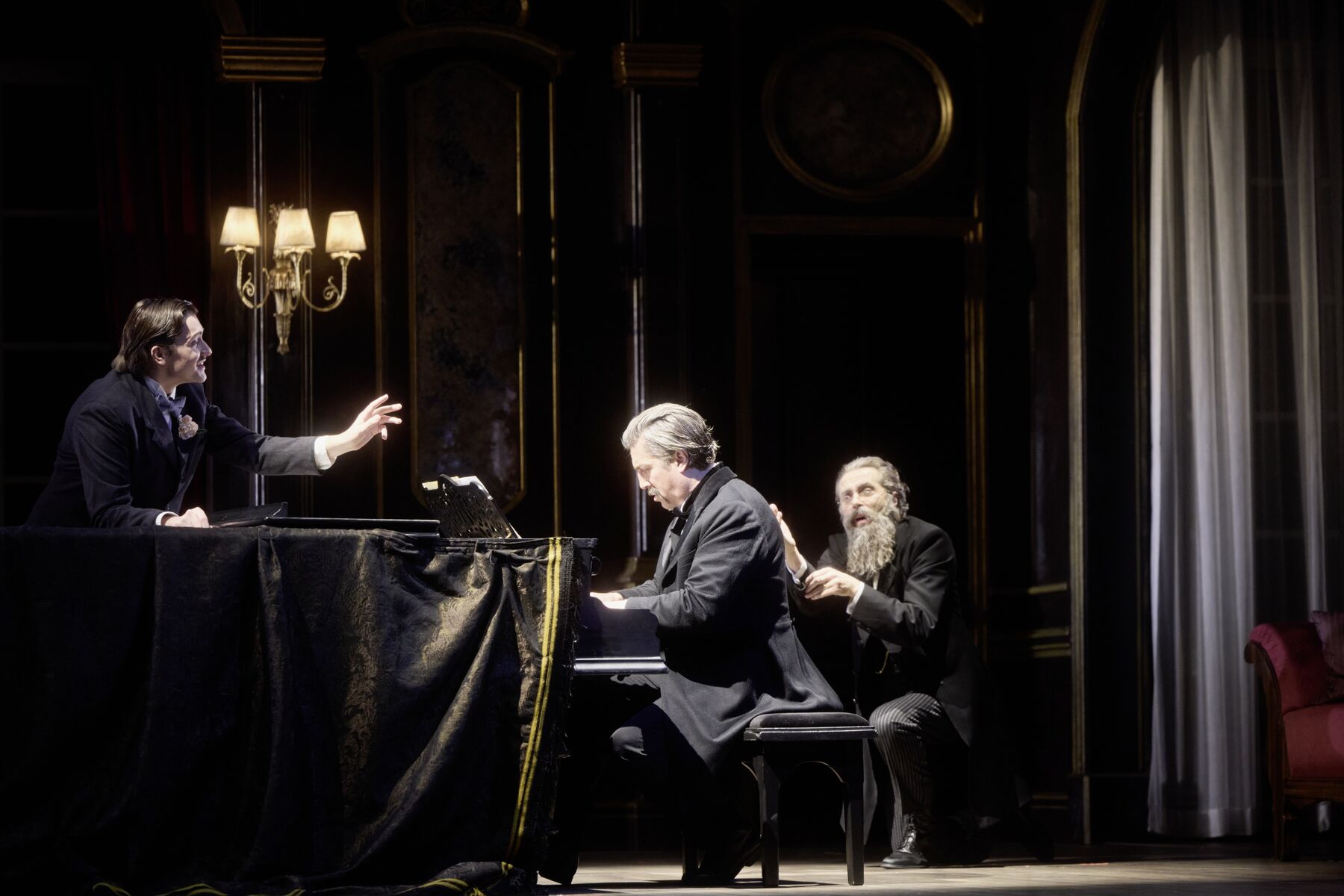 ein Mann in einem schwarzen Anzug spielt an einem Flügel. Links neben dem Flügel steht ein weitere Mann, der die Hand ausstreckt. Hinter dem Mann am Flügel sitzt ein weitere Mann der erschrocken schaut und einen langen Bart hat. 