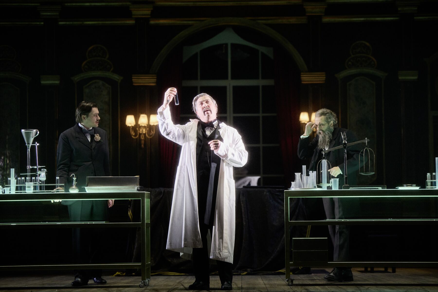 ein Mann mit einem weißen Laborkittel steht zwischen zwei Labortischen die vor einem großen prunkvollen Fenster stehen. Er schaut in die Ferne und hält dabei ein kleines Reagenzglas. Im Hintergrund stehen seitlich von ihm zwei weitere Männer, die sich gegenseitig anschauen.