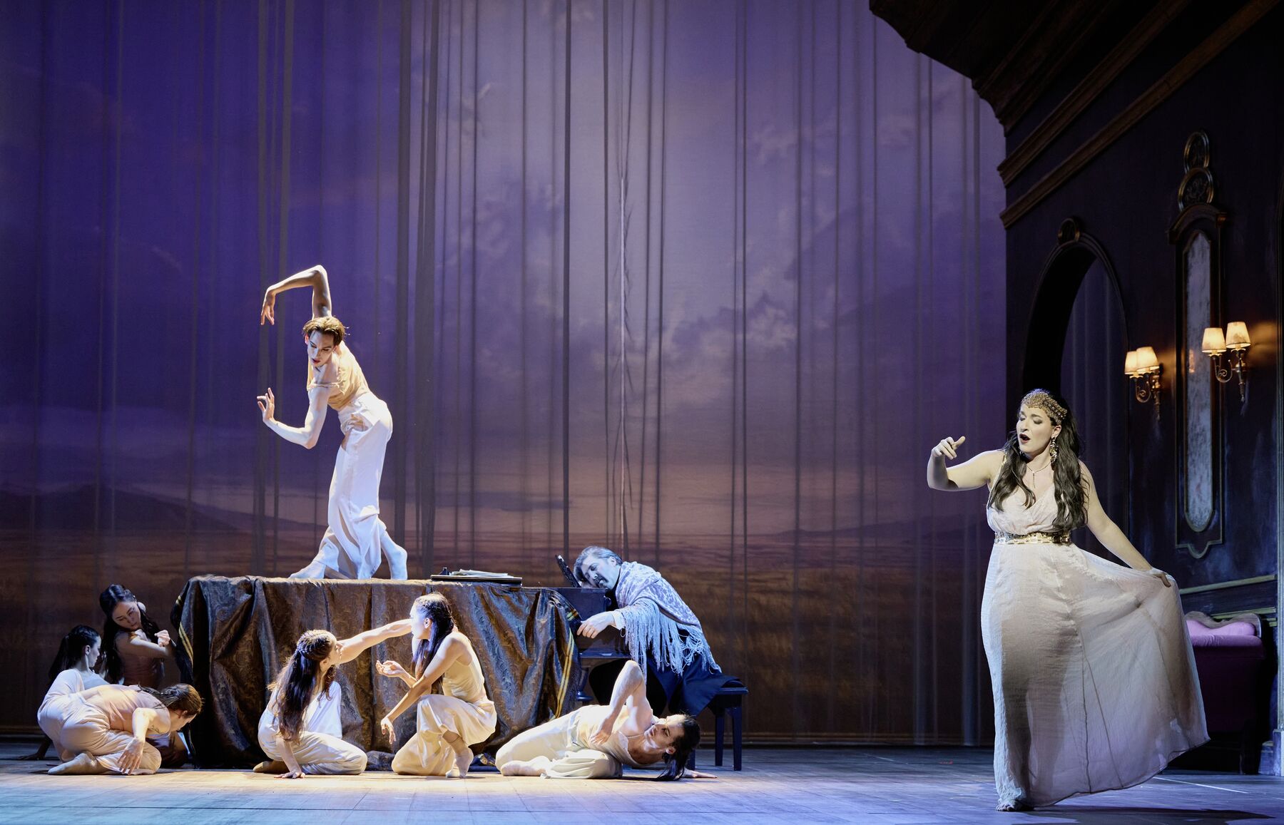 Vorne rechts steht eine Frau in einem bodenlangem weißen Kleid. weiter hinten sitzt ein Mann schlafend an einem Flügel. Um und auf dem  Flügel bewegen sich anmutig Tänzer in weißen Kostümen.