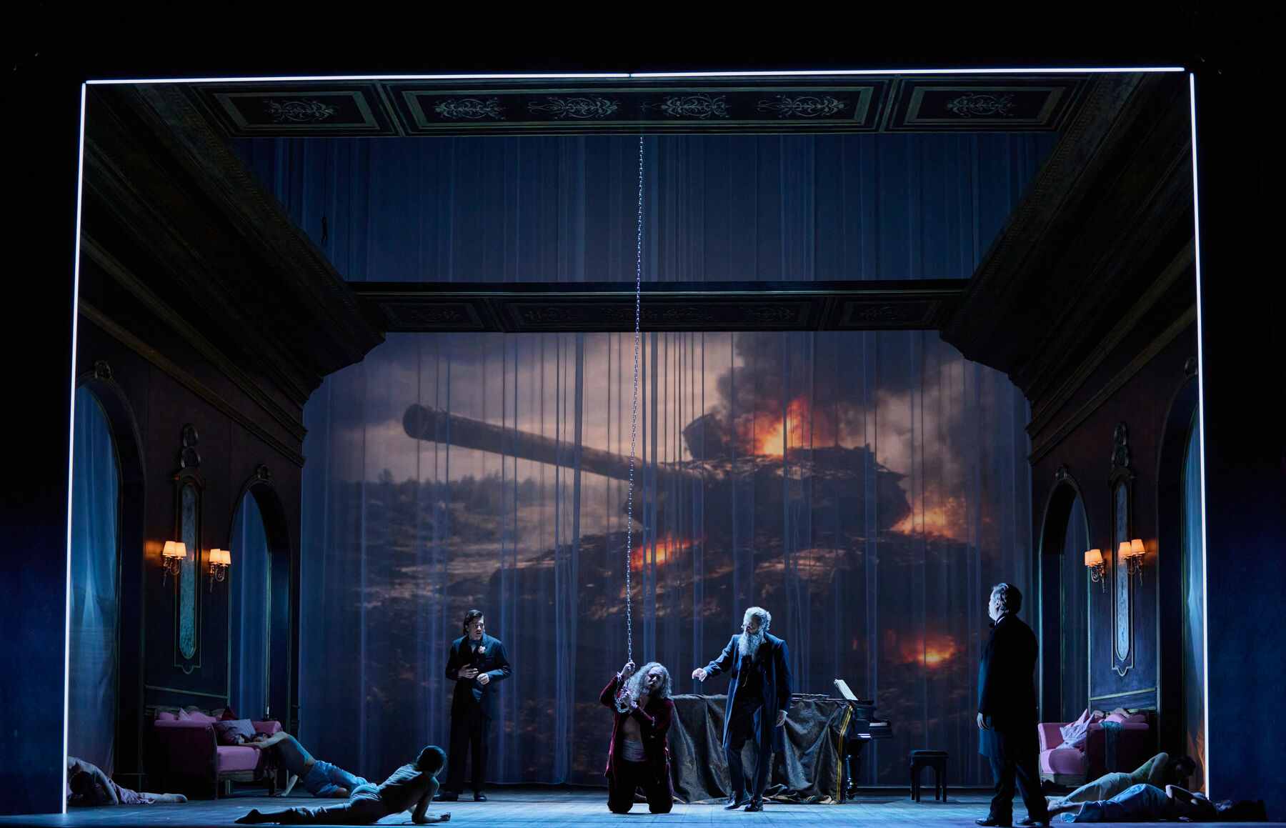 in einem prunkvollen Saal kniet ein Mann angekettet in der Mitte. Um ihn herum sind mehrere liegende Menschen und drei stehende Männer in schwarzen Anzügen. Im Hintergrund sieht man eine große Produktion eines brennenden Panzers.