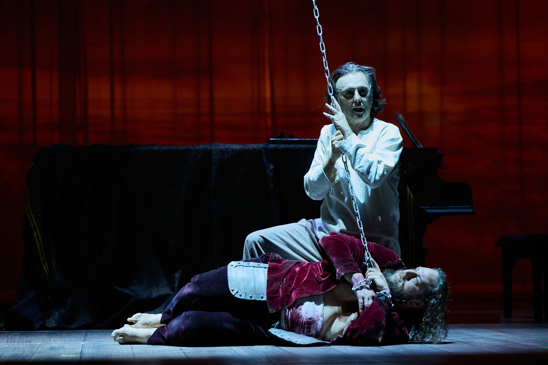 ein verwundeter Mann liegt am Boden. Er ist an eine Kette die von oben kommt gefesselt. Hinter ihm kniet ein Mann in weiß der die Kette festhält. Im Hintergrund steht ein Flügel.