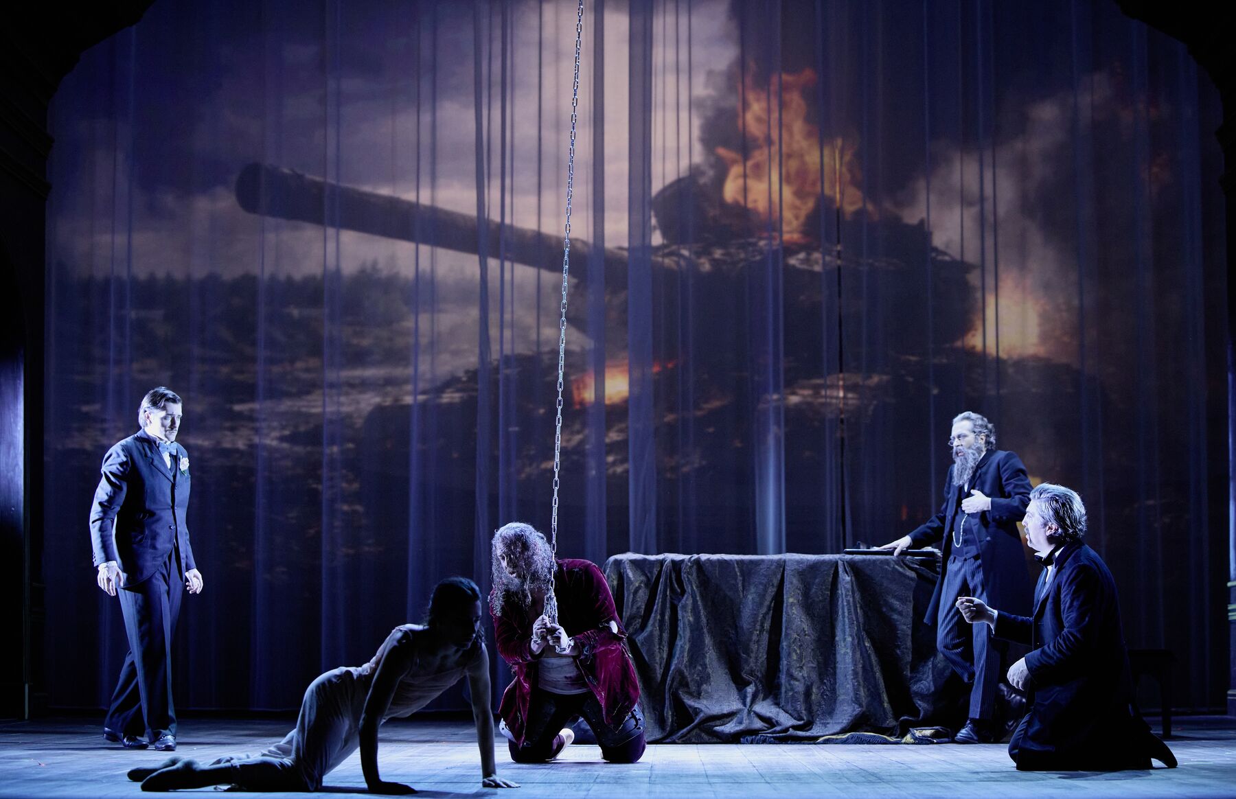 in einem prunkvollen Saal kniet ein Mann angekettet in der Mitte. Um ihn herum sind drei erschrockene Männer und eine sich aufstützende Tänzerin. Im Hintergrund sieht man einen Flügel und eine große Produktion eines brennenden Panzers.