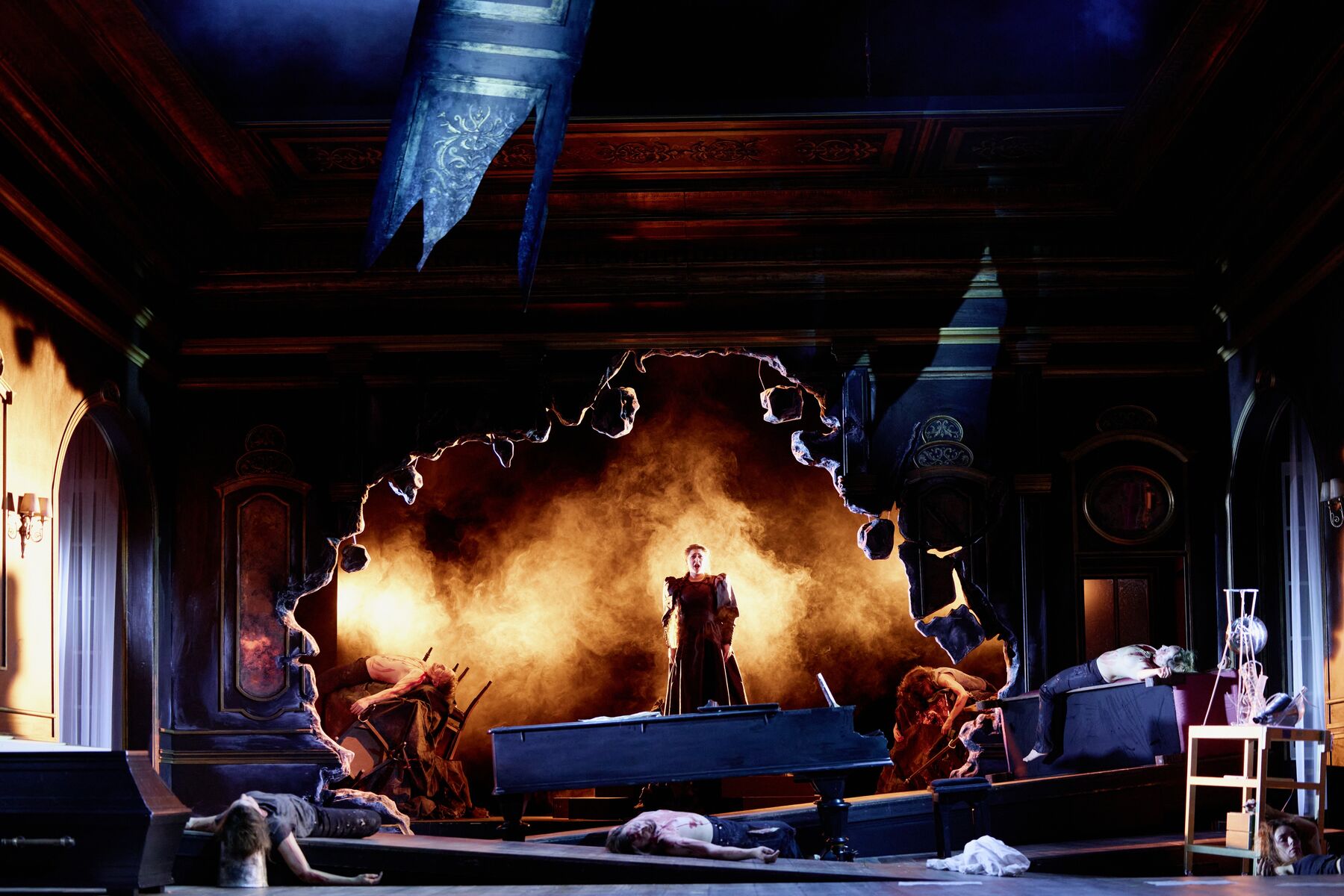 in einem zerbombten prunkvollen Saal liegen mehrere tote Menschen. In der Mitte vor einem riesigen zerbombten Loch steht auf einem Flügel eine Frau in einem pompösen Kleid.