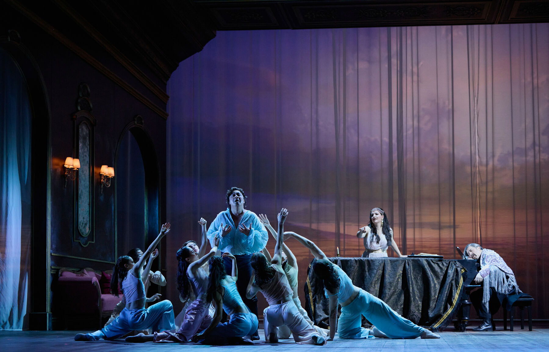 sieben Tänzer knien tanzend vor einem Mann der verzweifelt aussieht. Hinter ihm schläft ein Mann an einem Flügel, eine Frau lehnt sich über den Flügel und zeigt auf den verzweifelten Mann.