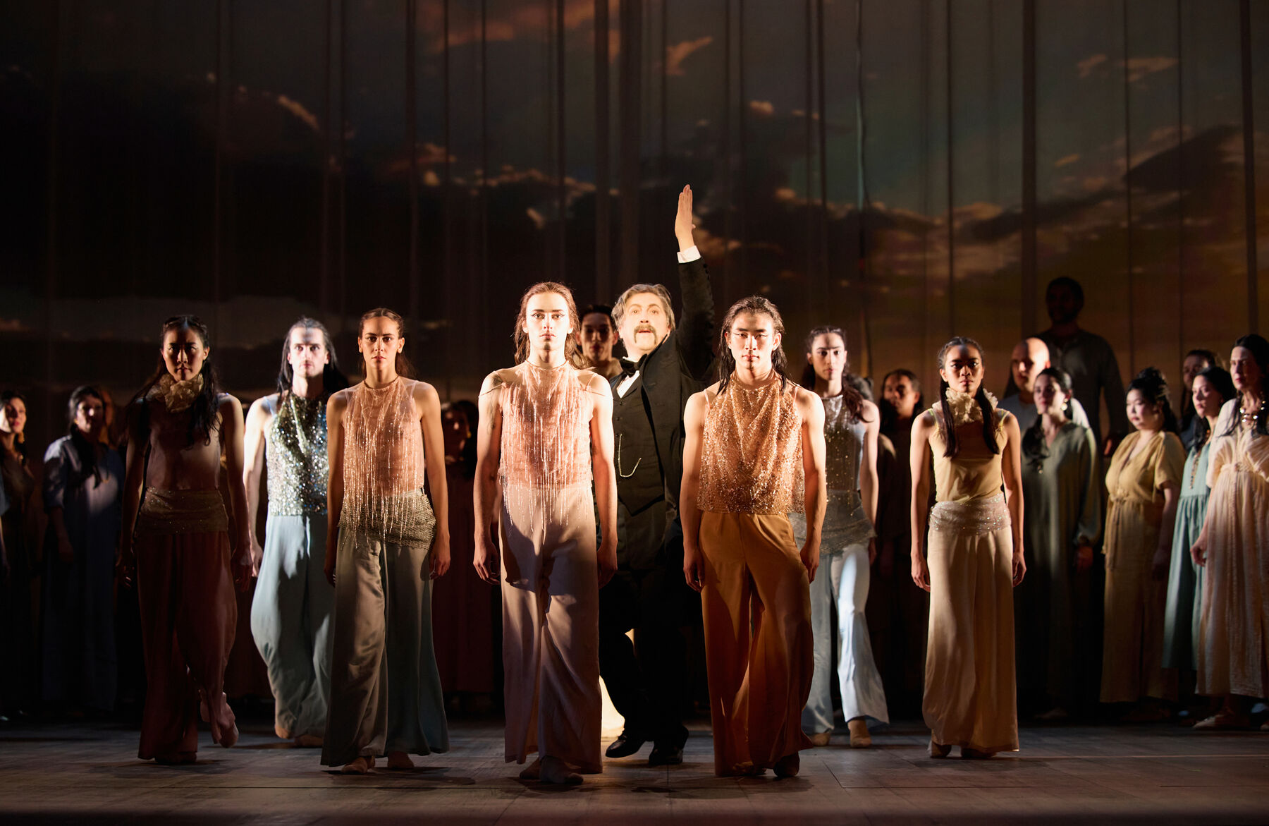 sieben Tänzer in glitzernden Kostümen stehen in einer V-Formation vor einem Mann in einem schwarzen Anzug welche nach ob zeigt. Hinter ihnen stehen viele Menschen in festlicher Kleidung.