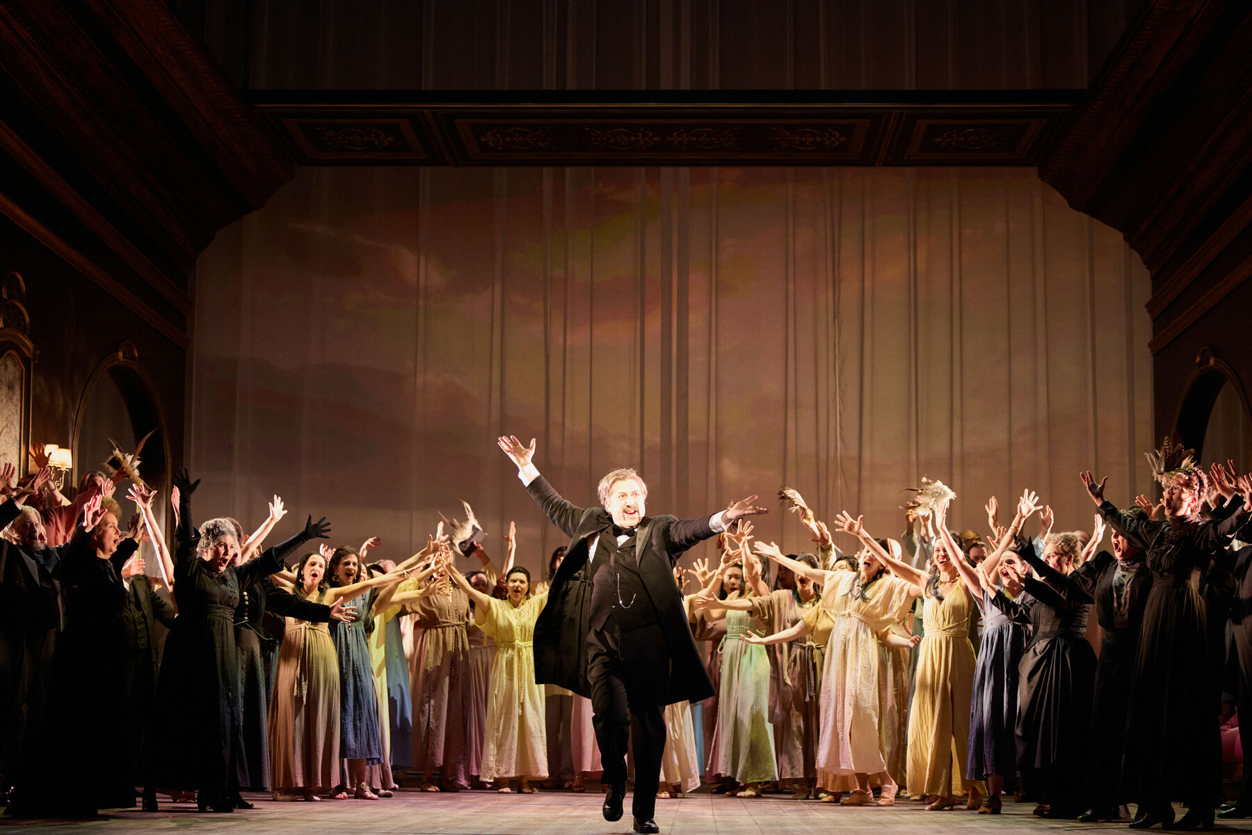 ein Mann in einem historischen schwarzen Anzug steht euphorisch mit erhobenen Händen in der Mitte von vielen euphorischen Menschen in festlicher Kleidung.