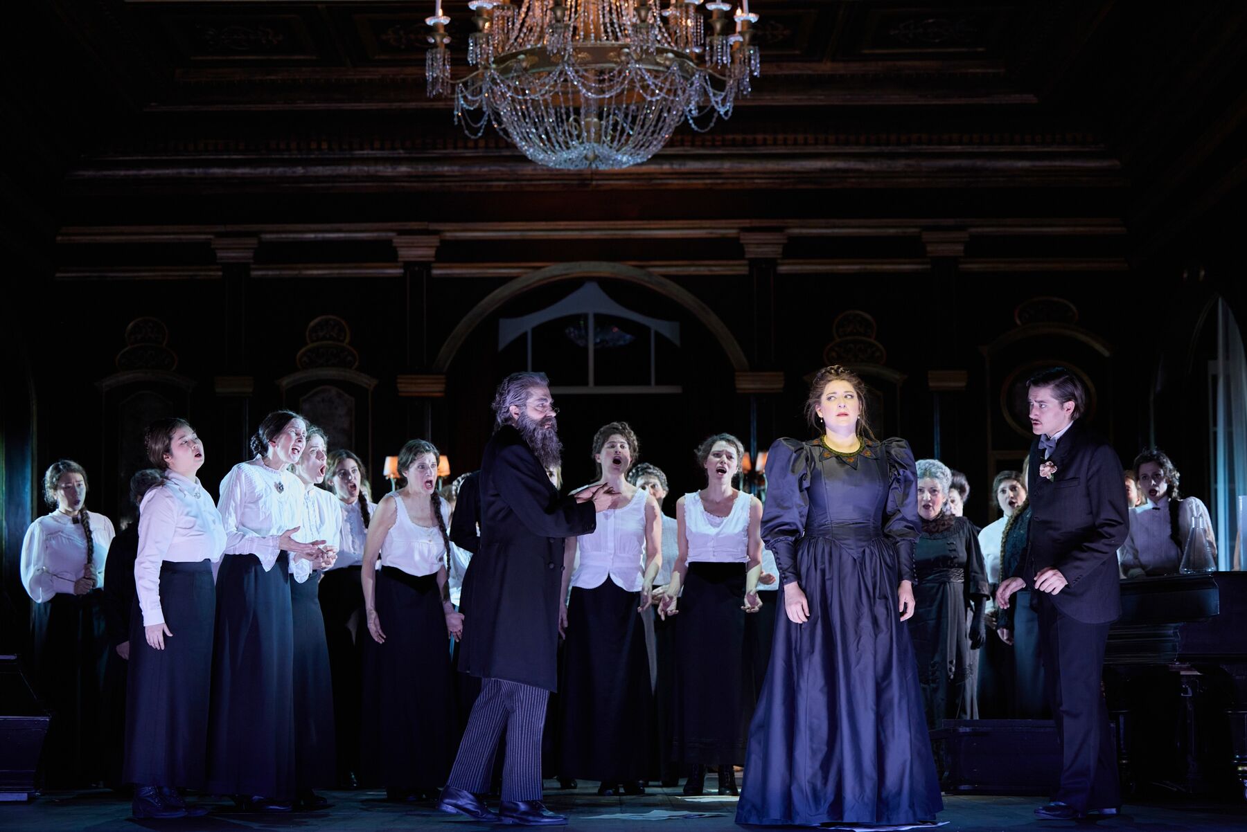 eine Frau in einem pompösen schwarzen Kleid steht vor mehreren Frauen in schwarz weißen Kleidern. Neben ihr stehen zwei  zu ihr gerichtete Männer in schwarzen Anzügen.