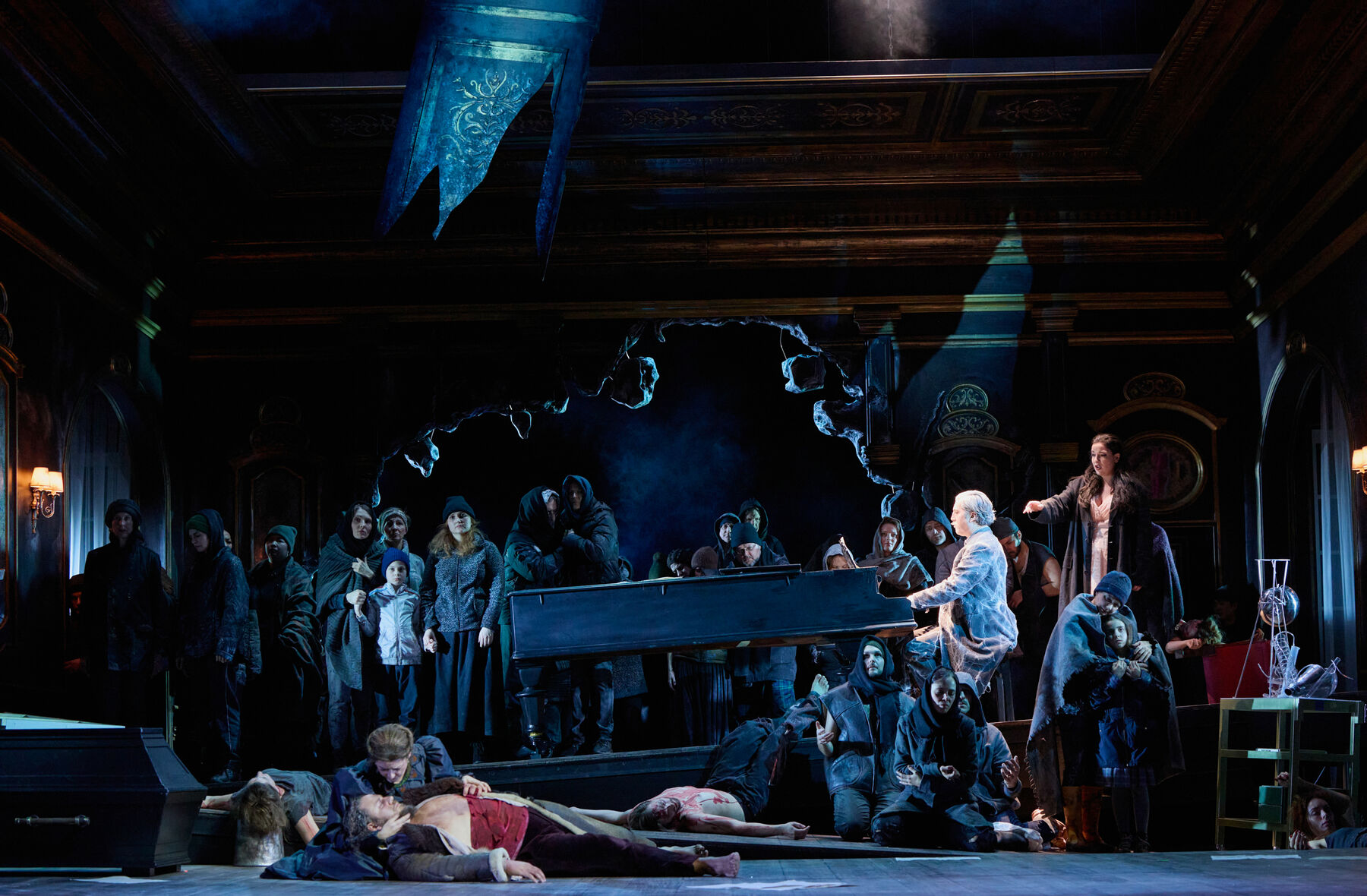 in einem zerbombten Saal stehen viele Menschen in moderner Kleidung. vor ihnen steht eine Flügel an dem ein Mann wie eine Statue sitzt. Ganz vorne hält eine kniende Frau in einem schwarzen Kleid einen sterbenden Mann.
