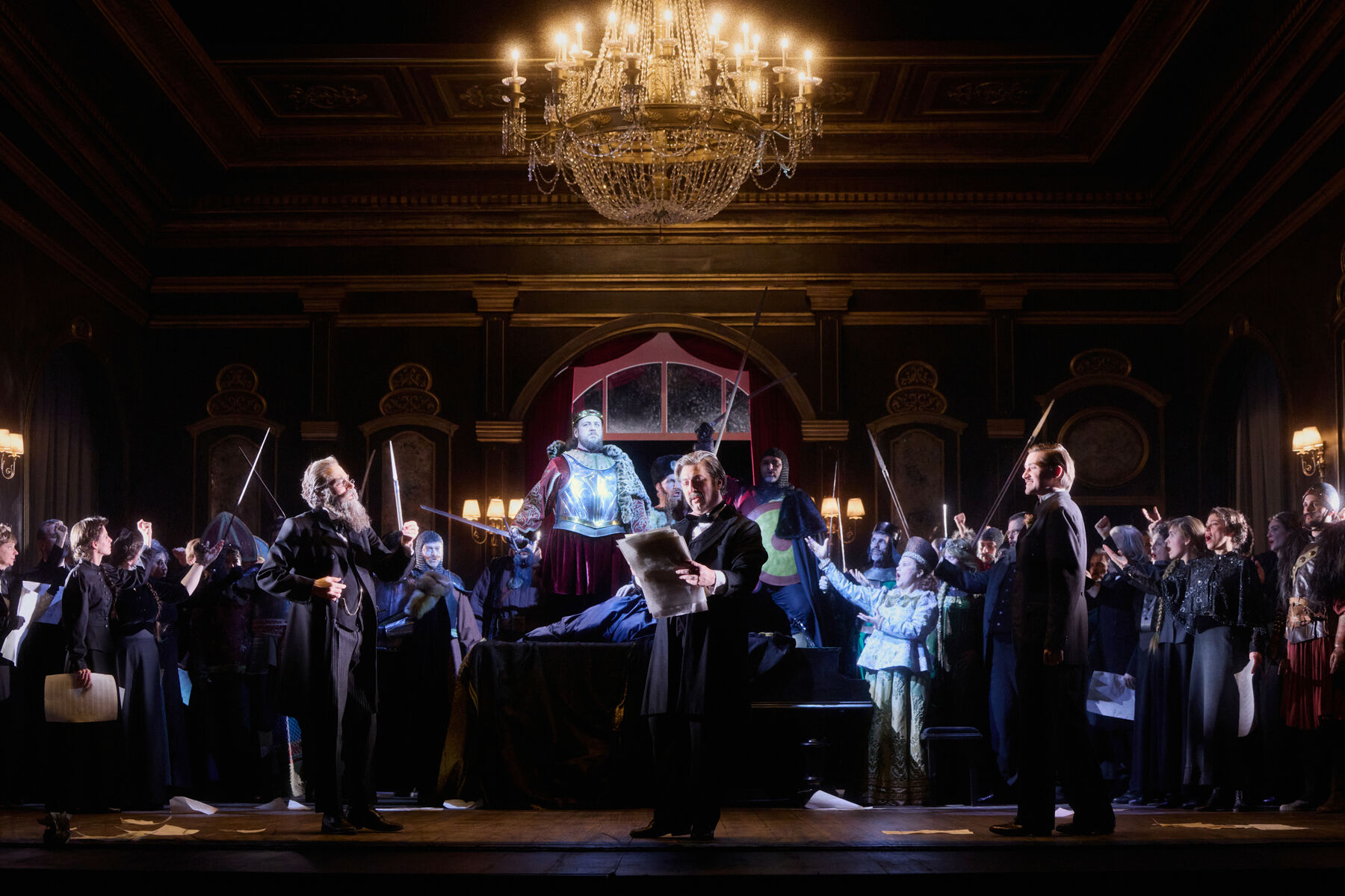 viele Menschen in historischen Kostümen stehen in einem prunkvollen Saal. Einige von ihnen heben Schwerter in die Luft. In der Mitte steht erhöht ein Krieger mit einem Schwert.
Im Vordergrund stehen drei Männer mit schwarzen Anzügen. Der Mittlere hält viele Papiere in der Hand.