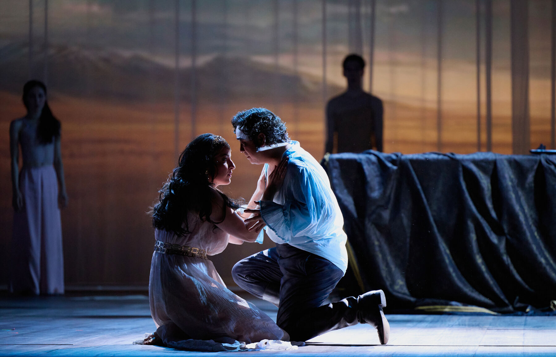 eine Frau in einem weißen Kleid und ein Mann mit einem weißen Hemd knien auf dem Boden und halten sich gegenseitig. Im Hintergrund sieht man eine Landschaft und zwei Menschen die davor stehen.