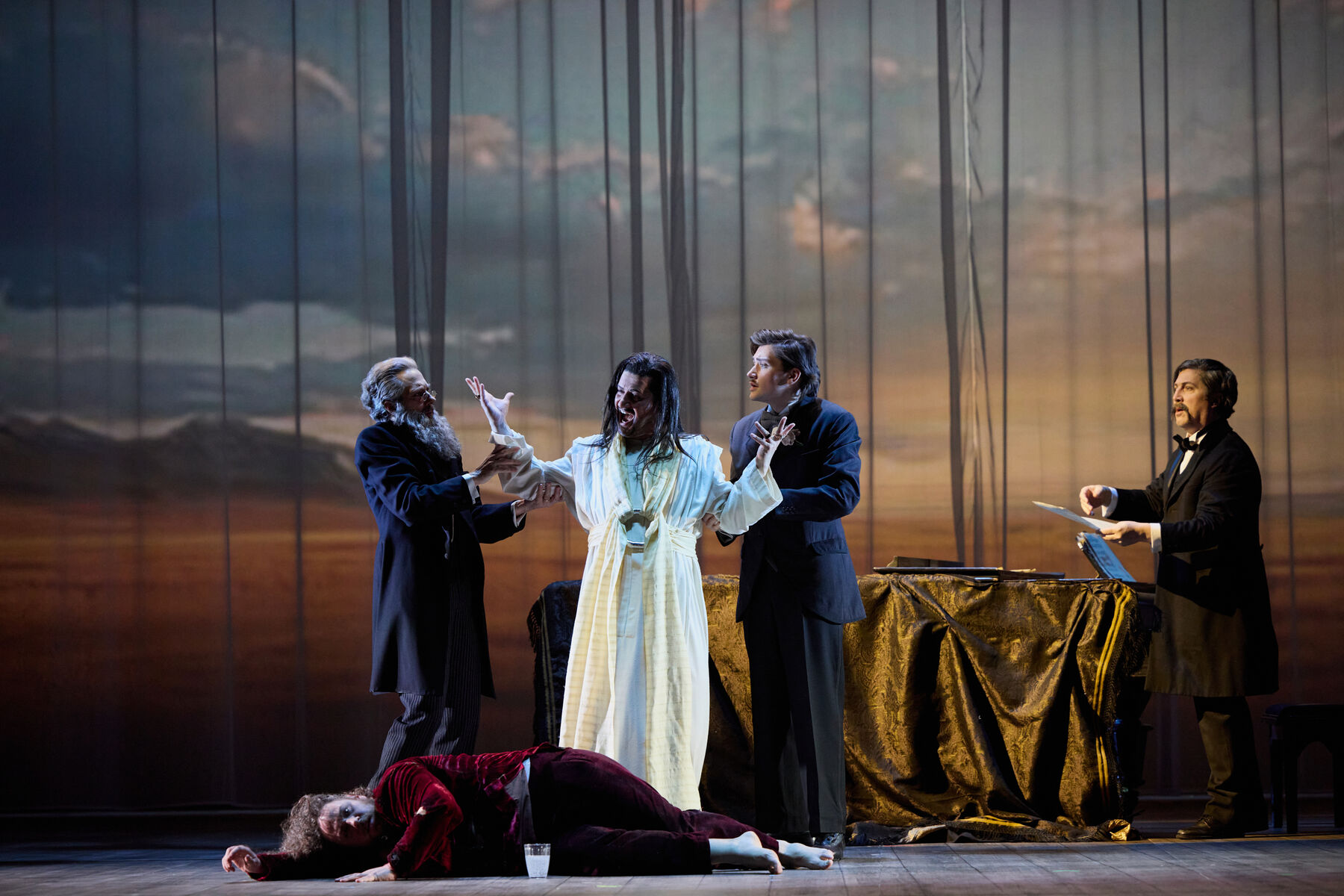 ein schreiender Mann in einem weißen Gewand wird von zwei Männern in schwarzen Anzügen festgehalten. Vor ihnen liegt ein Mann in roter Kleidung. Im Hintergrund steht ein Mann an einem Flügel.