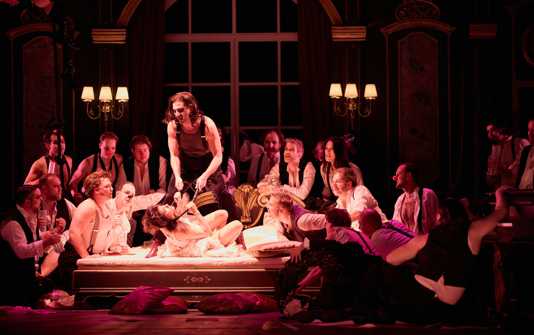 eine leichtbekleidete Frau liegt auf einem Bett umgeben von mehreren Männern. Ein Mann Steht über ihr auf dem Bett und zieht sie mit einer Peitsche Am Hals zu sich hoch.