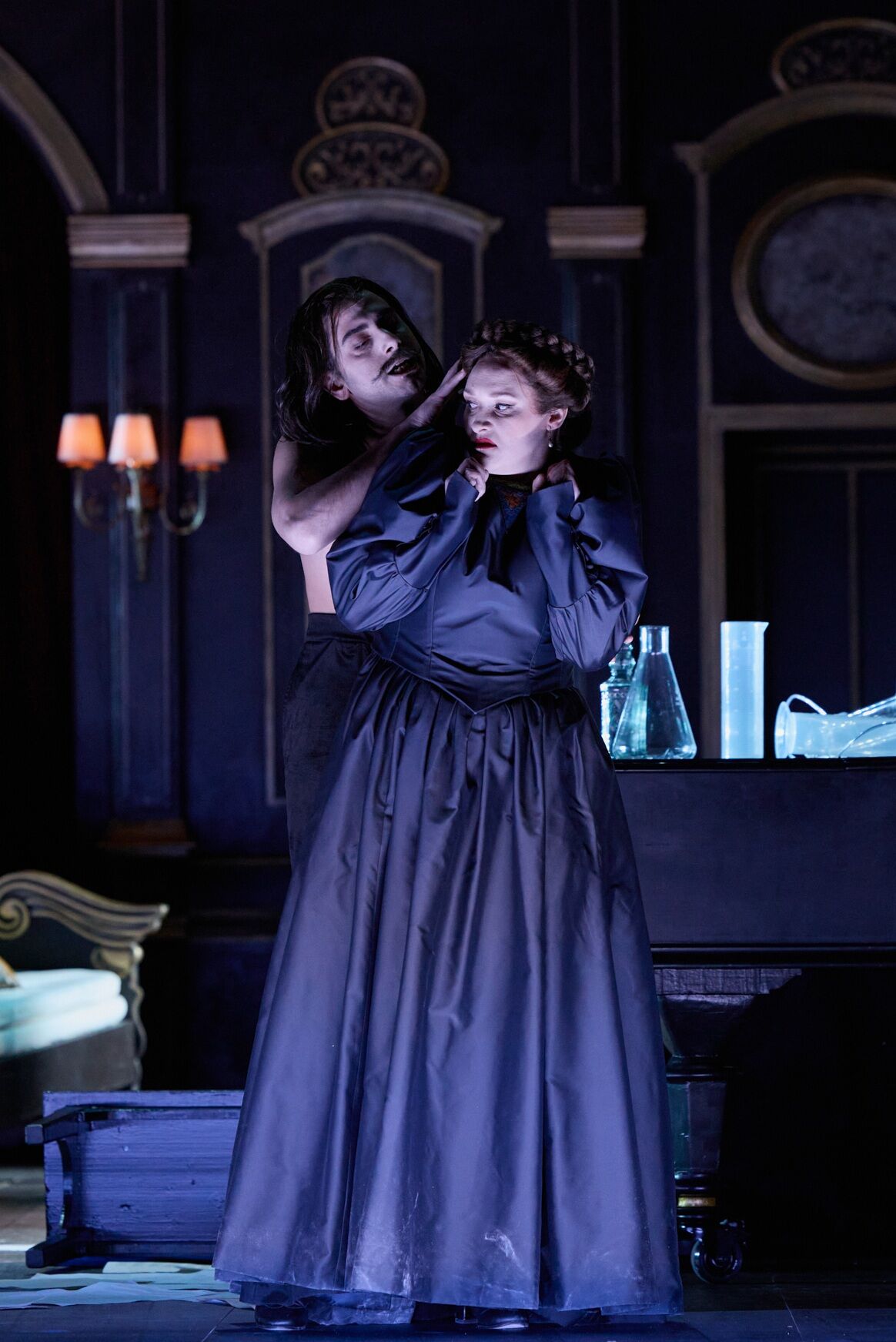 eine Frau in einem pompösen schwarzen Kleid steht vor einem oberkörperfreien Mann. Der Mann berührt ihre Haare und sie schreckt davor zurück.