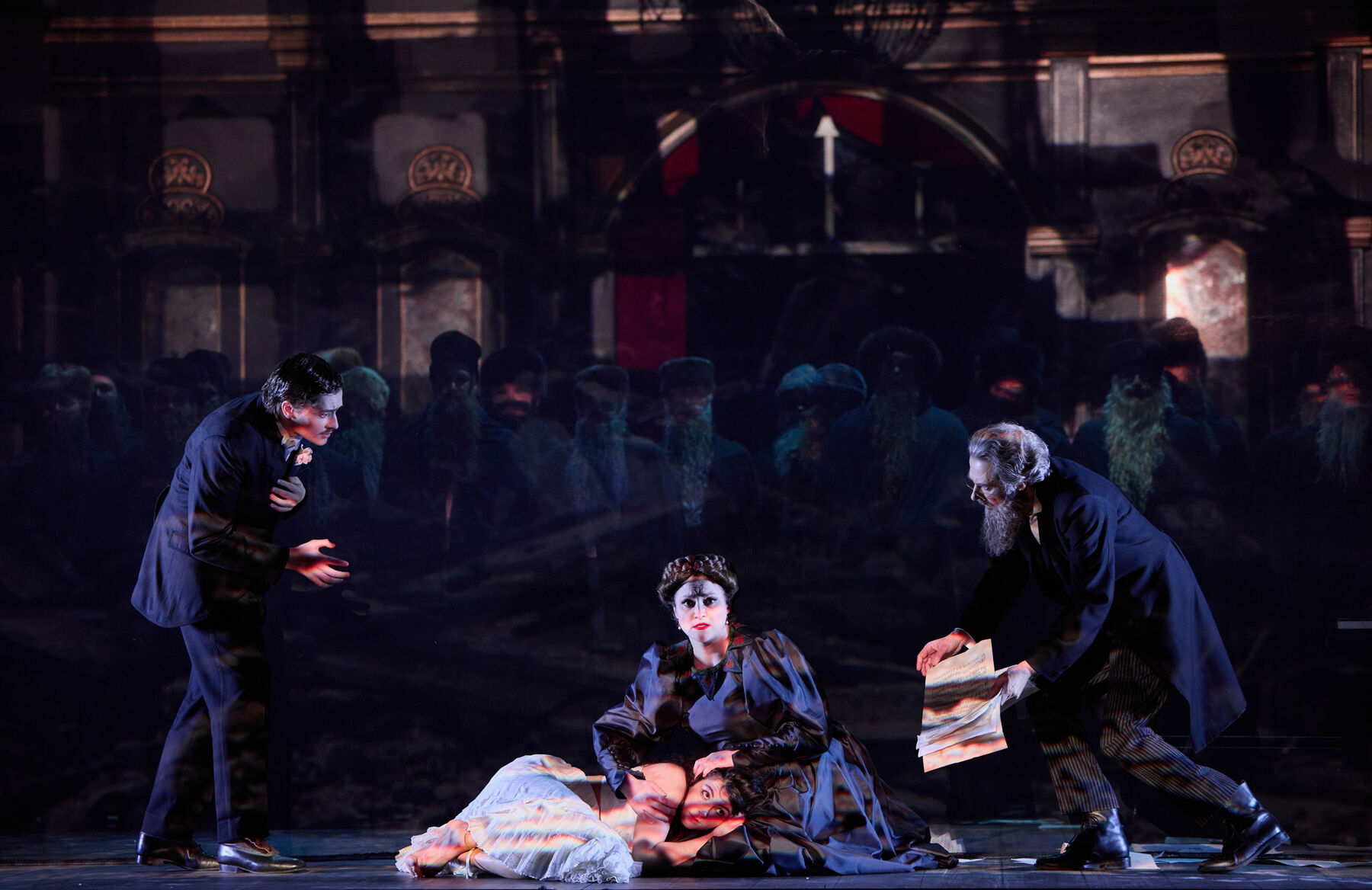 eine Frau in einem pompösen schwarzen Kleid kniet zwischen zwei Männern die sich zu ihr runterbeugen. In dem Schoß der Frau liegt eine zusammengekauerte, ängstliche Frau in einem weißen Kleid.